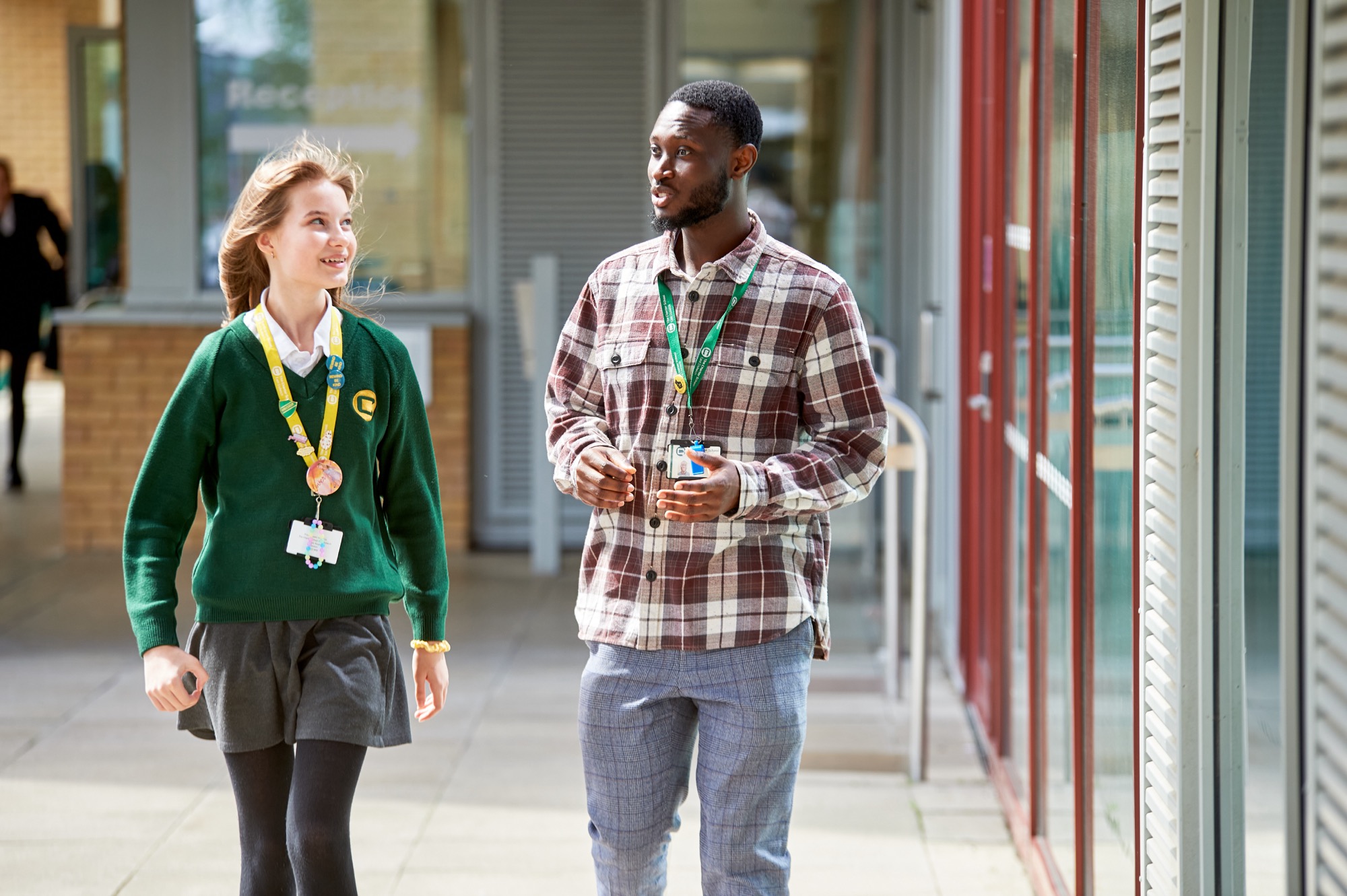 A young female student in a green sweater and gray skirt walks and smiles while talking with a young male teacher wearing a plaid shirt and blue pants in a school corridor.