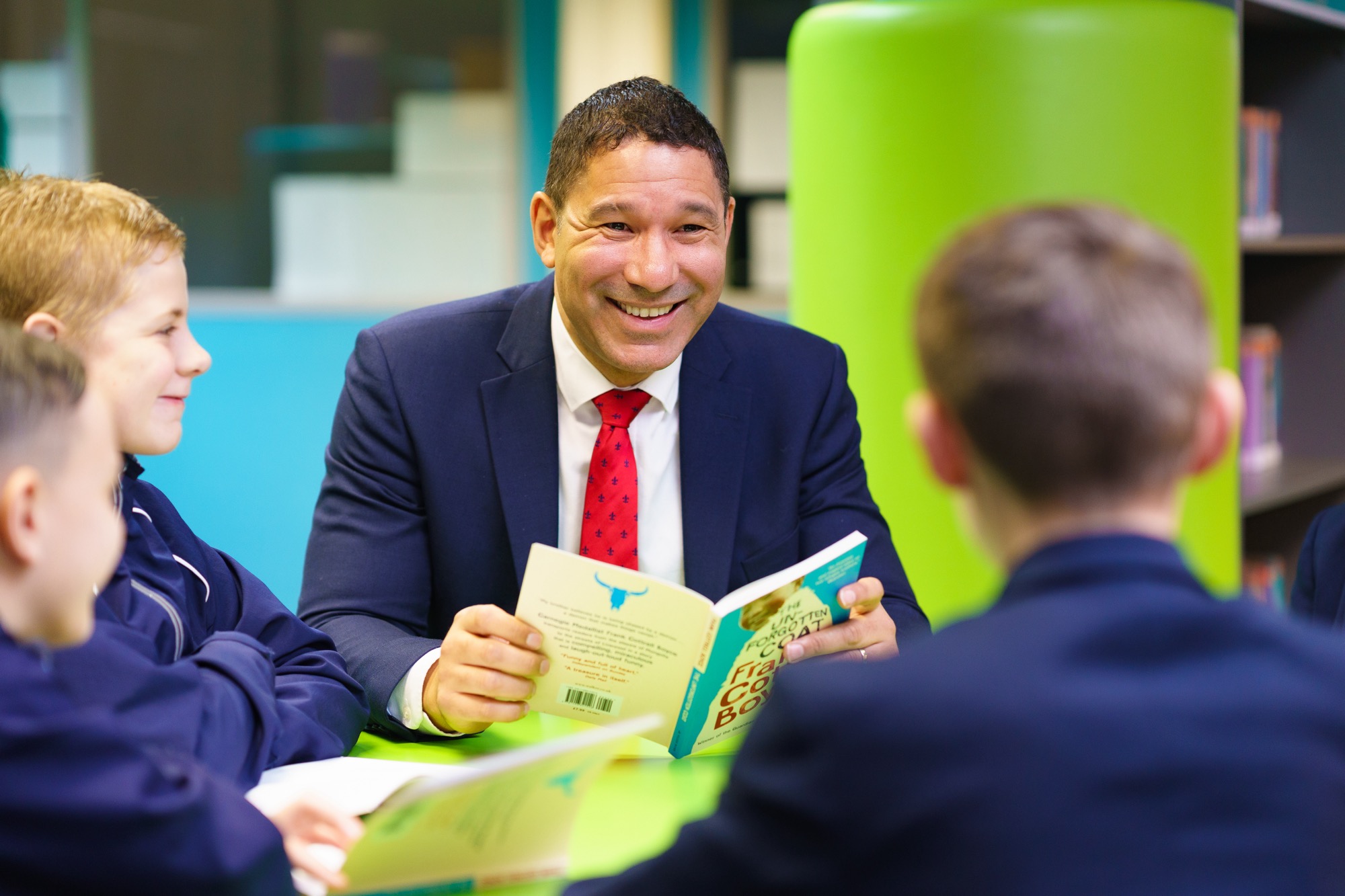 A man in a blue suit and red tie reads a book aloud to three boys in school uniforms sitting around a green table in a colorful school library.