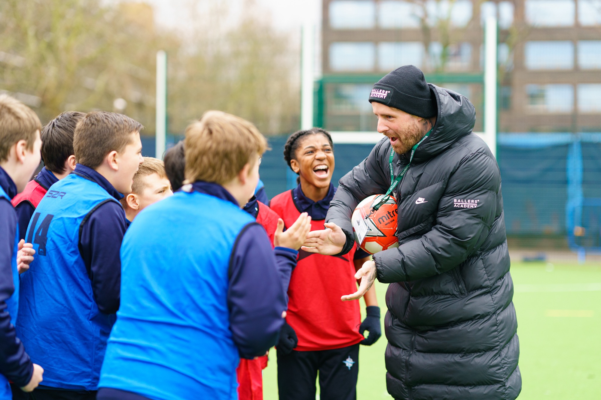 A coach wearing a black beanie and puffer jacket animatedly talks to a group of young soccer players on an outdoor field, holding a soccer ball and engaging with the kids who wear red and blue training vests, some smiling and clapping.