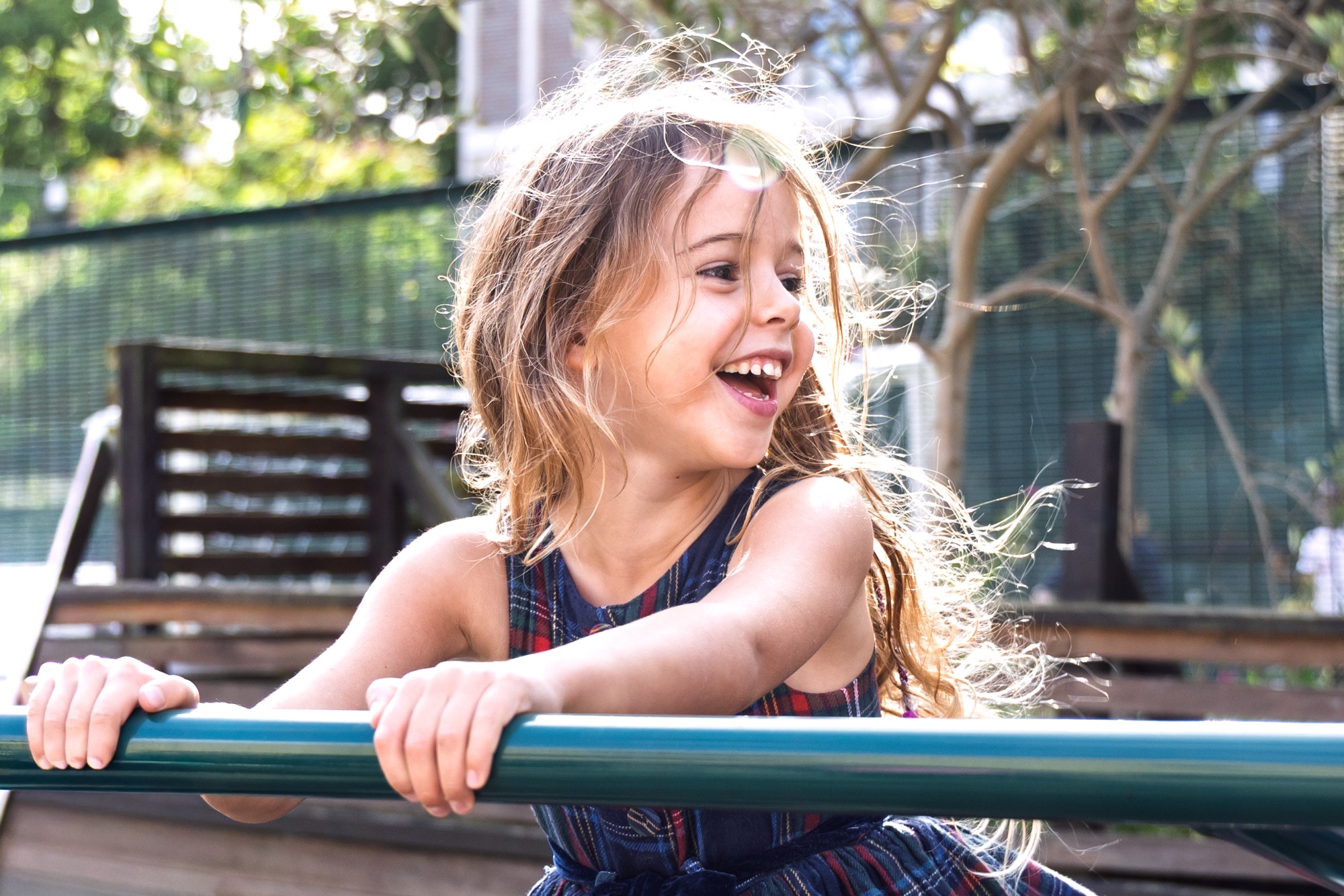 Smiling young girl with long hair holding onto a blue railing outdoors, with sunlight highlighting her hair and a blurred green fence and trees in the background.