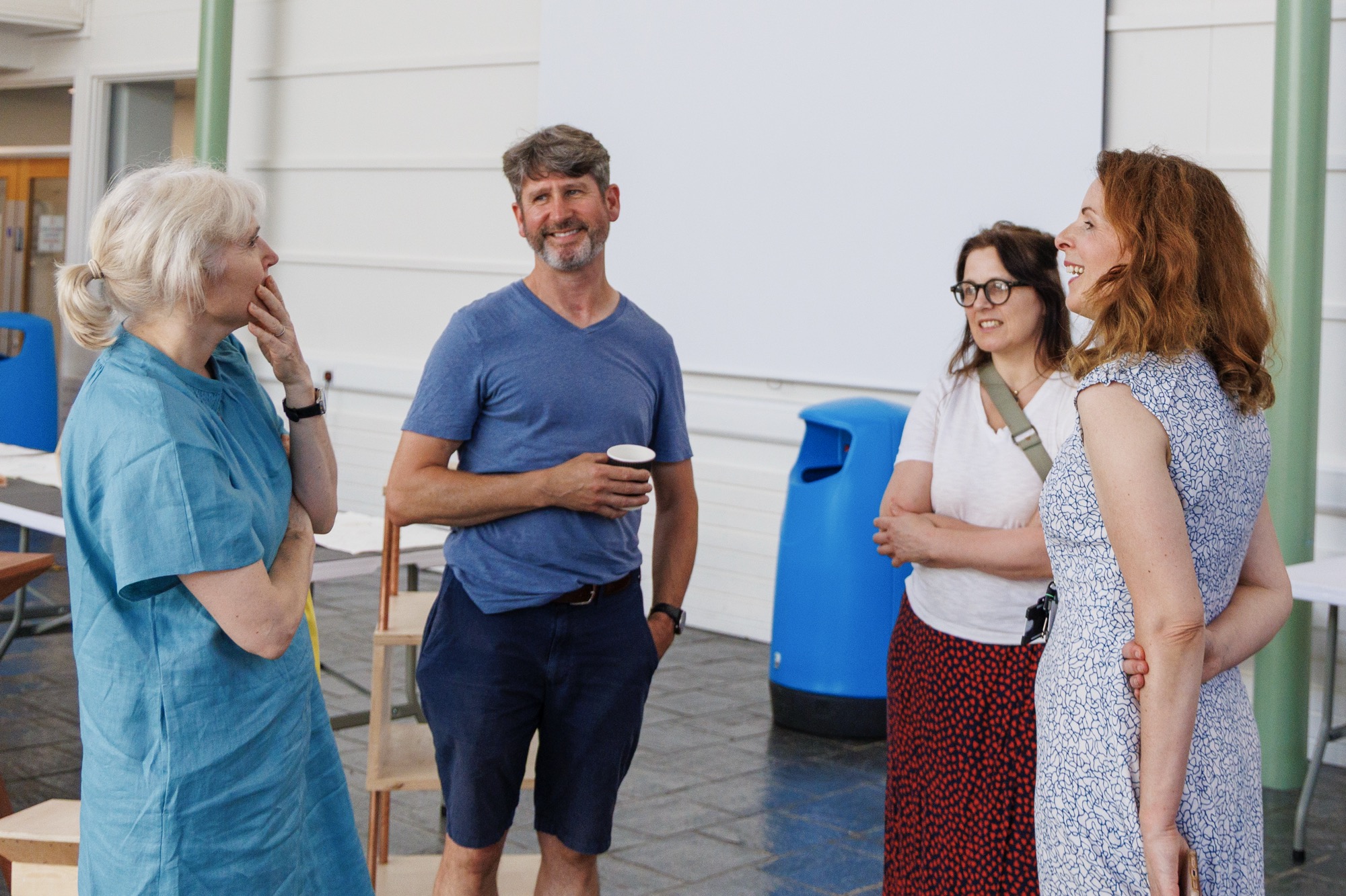 Four adults standing indoors in a casual setting engaged in conversation, with one man holding a paper cup, two women smiling, and one woman with hand on chin.