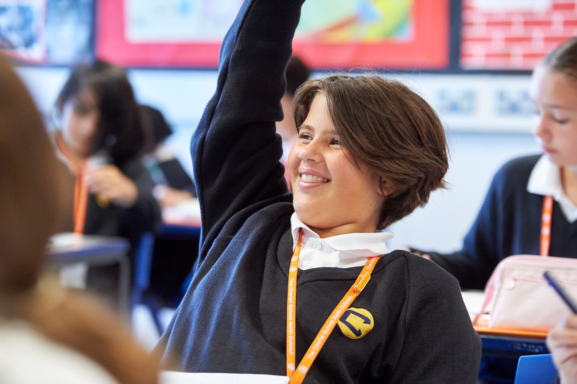 A smiling student in a dark sweater with a white collar raises their hand in a classroom while other students focus on their work.