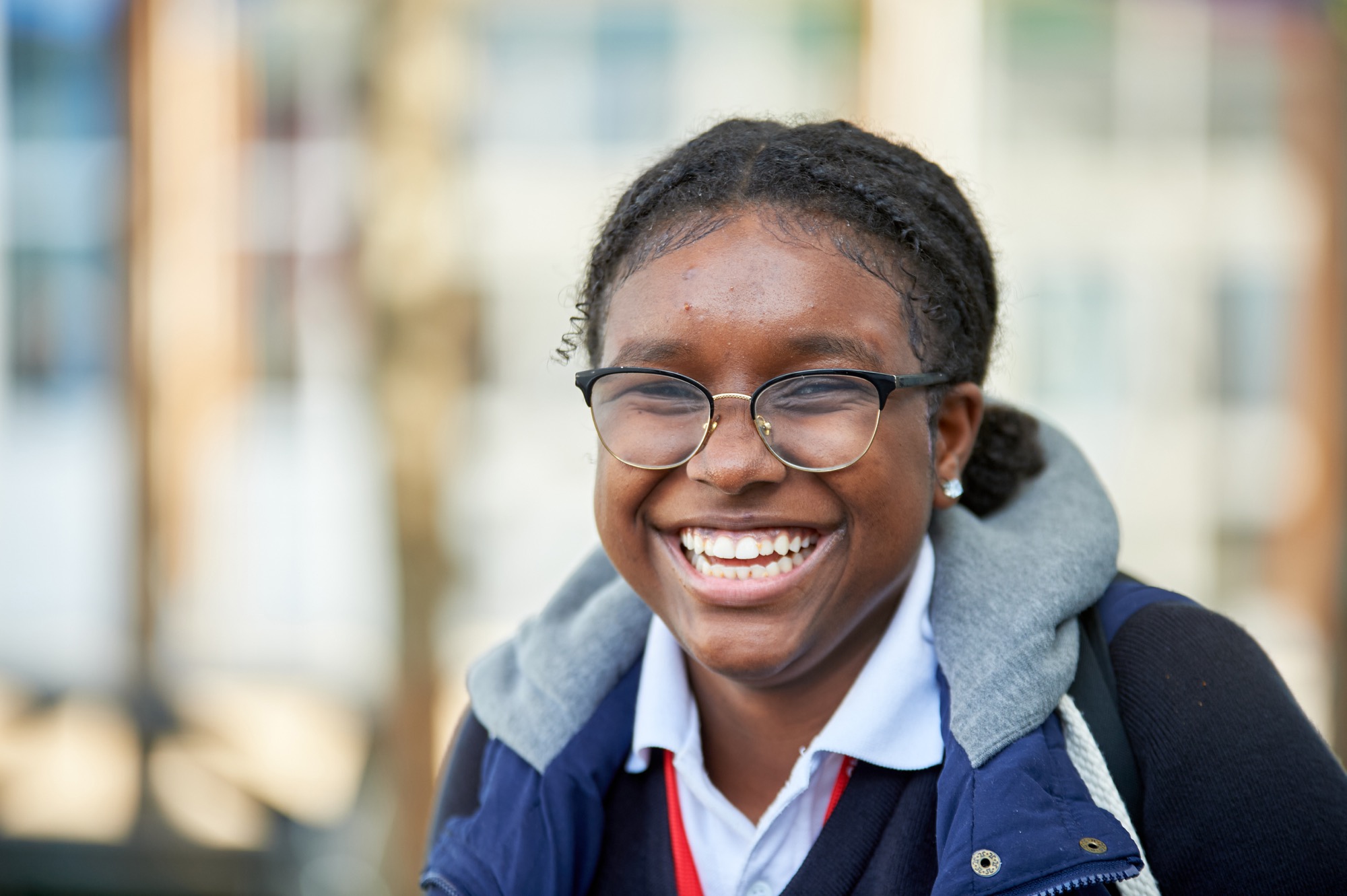 Smiling young person with glasses, wearing a white collared shirt, navy sweater, and blue jacket with a gray hood, standing outdoors.