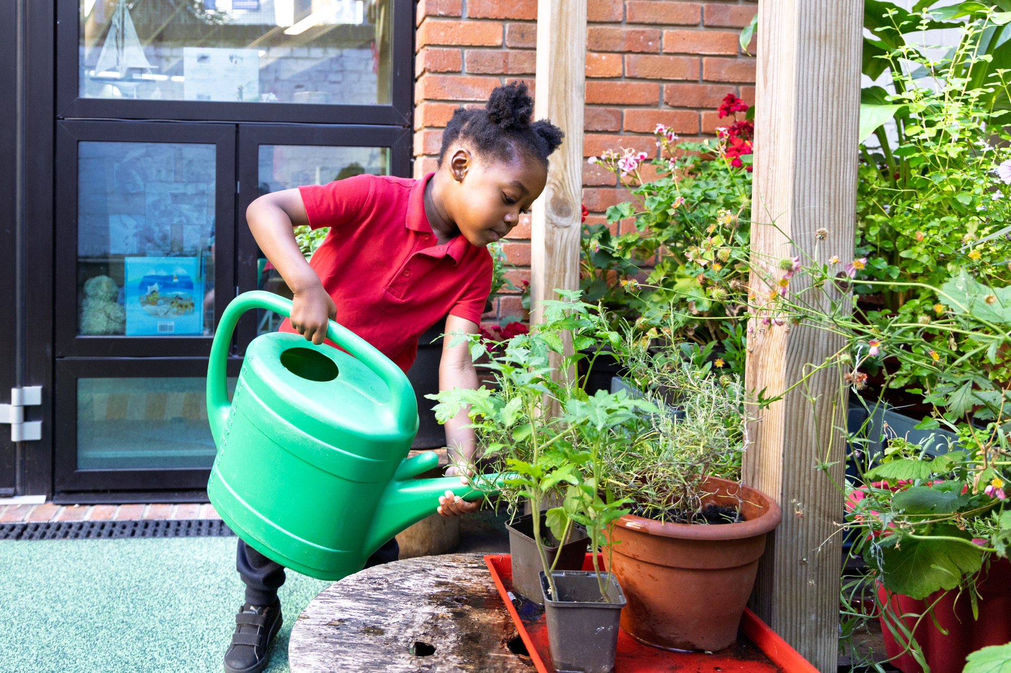 A young child in a red shirt waters plants in pots using a large green watering can, standing on a patio near a glass door and brick wall.