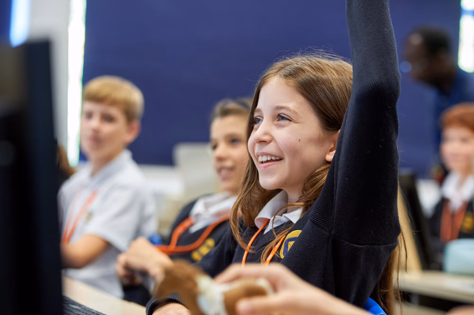 A smiling schoolgirl in a dark sweater raises her hand eagerly in a classroom with other attentive students.