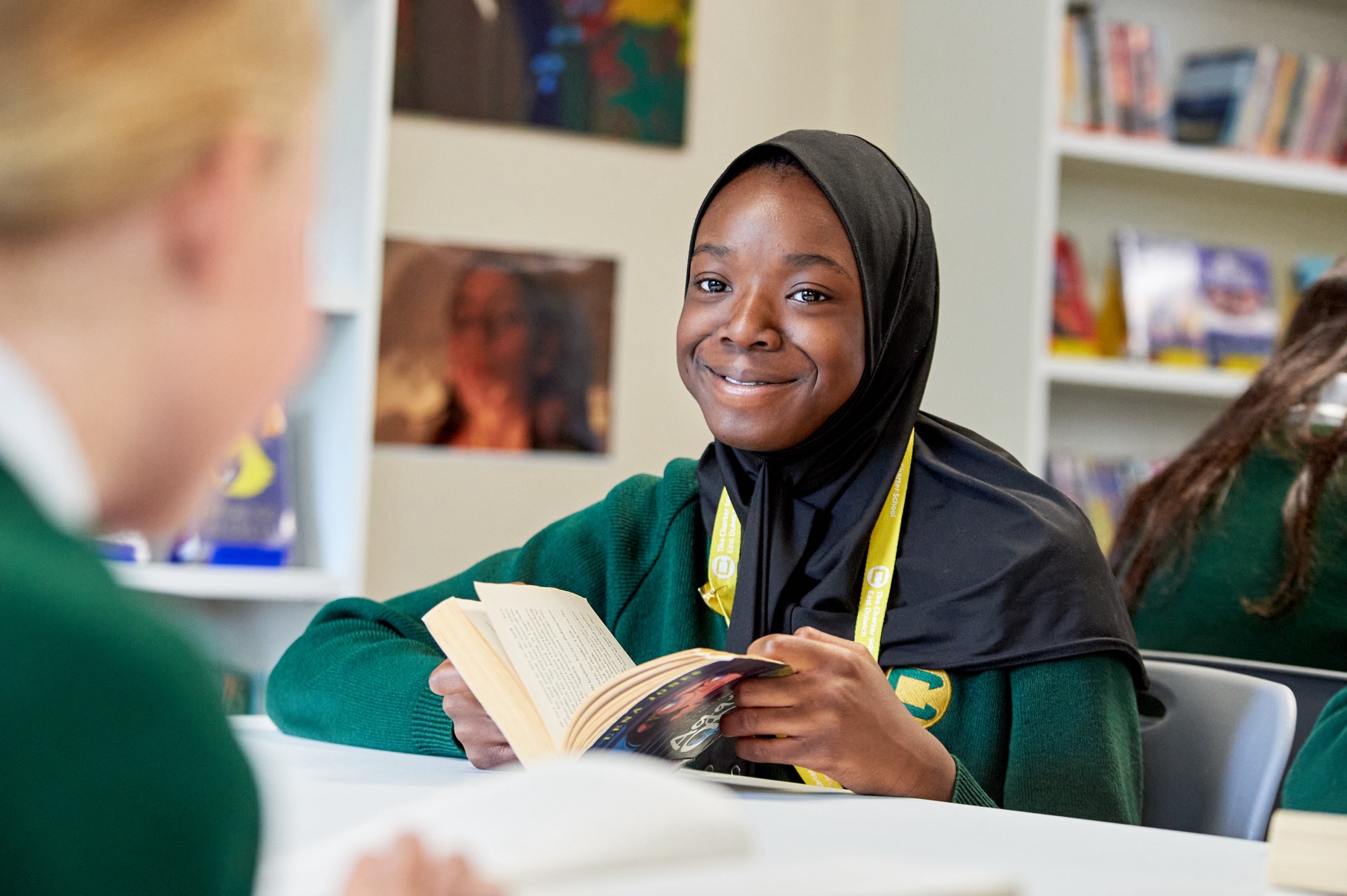 A smiling student wearing a black hijab and green sweater, holding an open book in a classroom or library setting with bookshelves in the background.