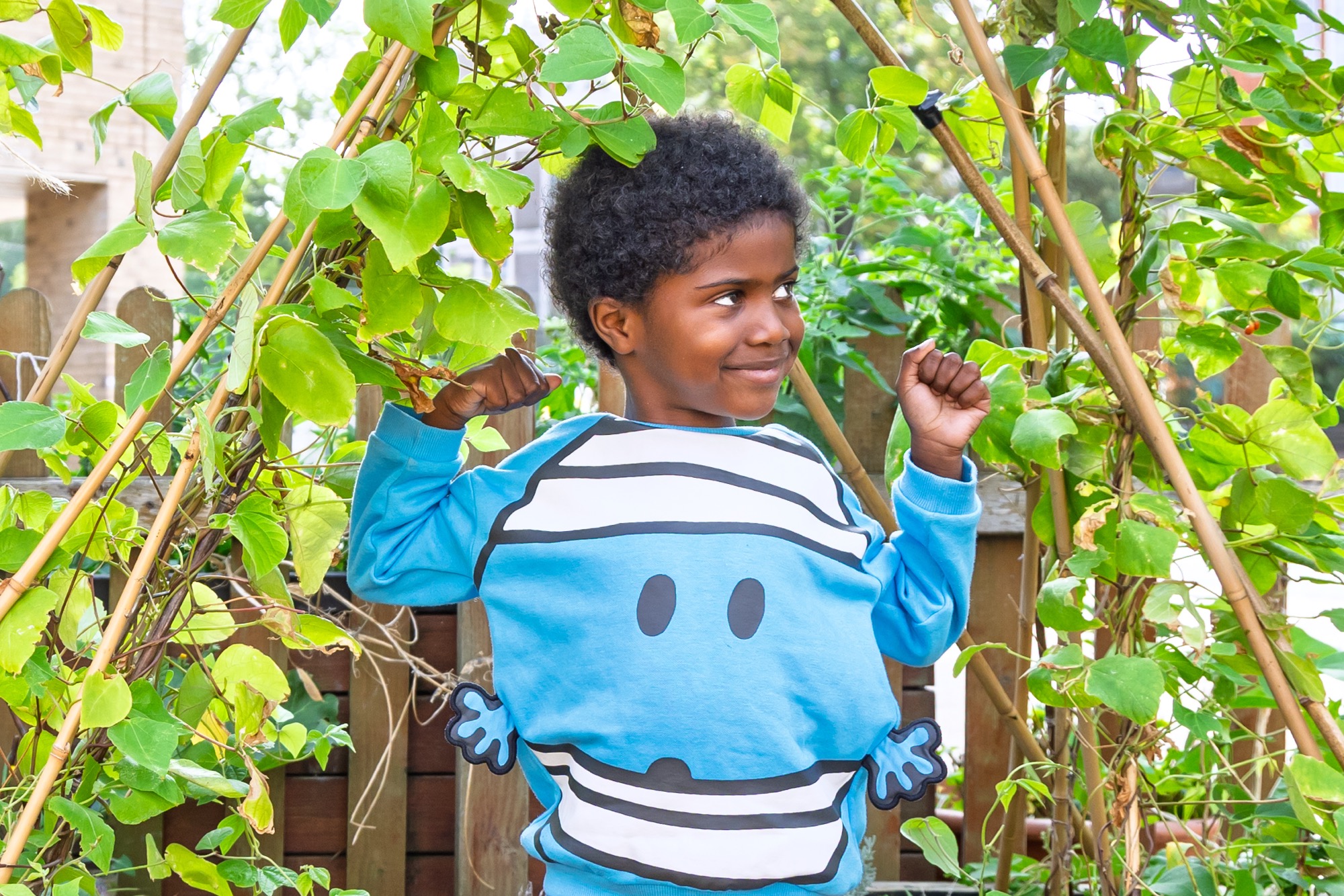 Smiling young boy wearing a blue sweater with a cartoon face, standing outdoors among green leafy plants and wooden fence, flexing his arms in a playful pose.