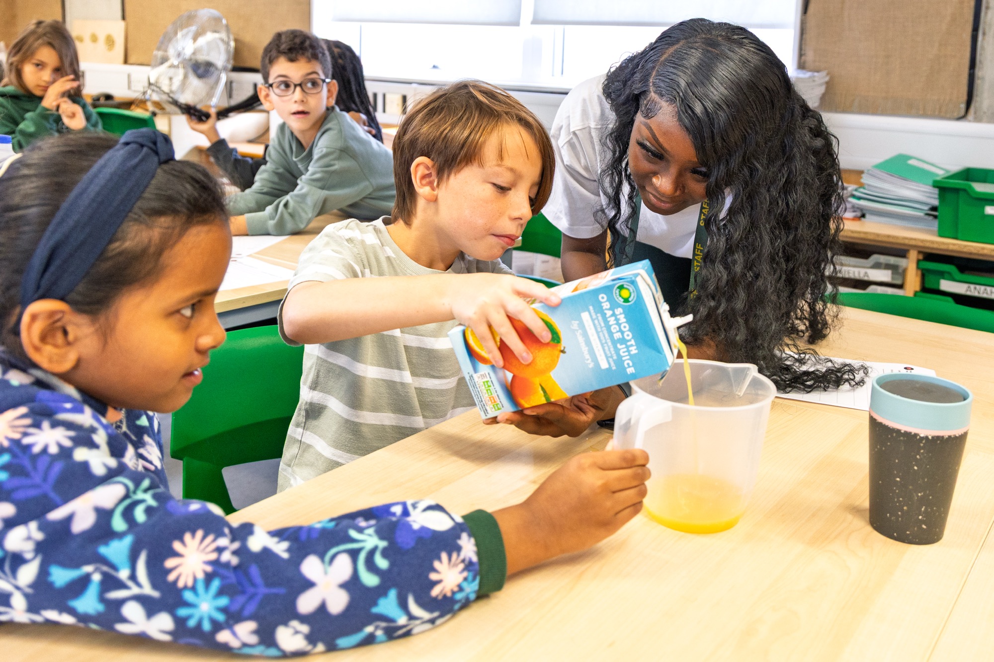 A boy in a striped shirt pours orange juice from a carton into a plastic measuring jug while a teacher with long black curly hair watches and a girl in a floral jacket holds the jug. They are sitting at a classroom table with other children and educational materials in the background.