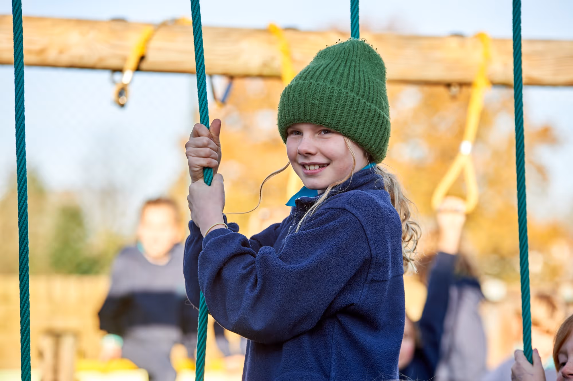 Smiling child wearing a green knit hat and navy blue jacket holding onto green ropes on a playground climbing structure with blurred children and autumn trees in the background.