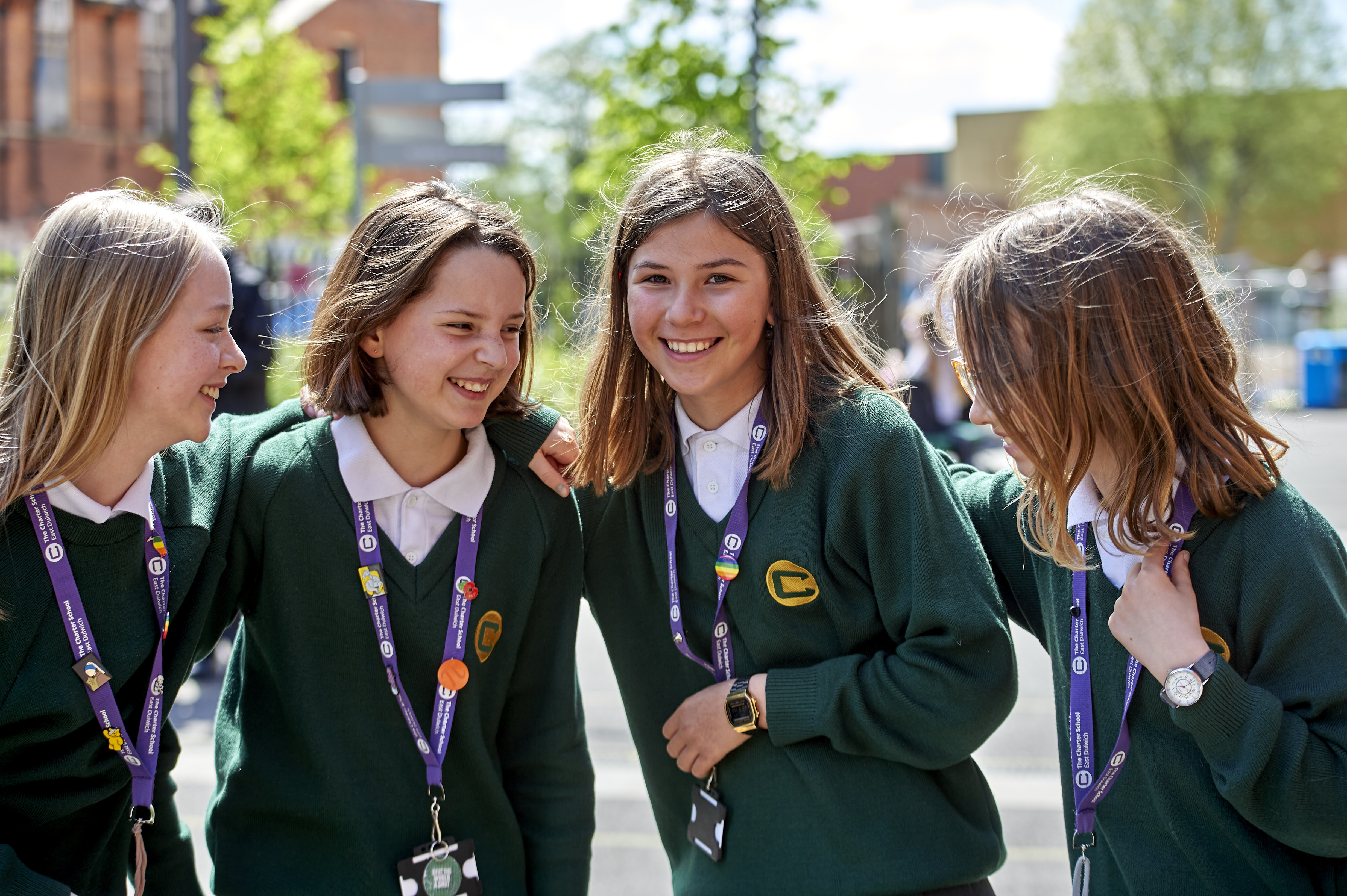 Four smiling teenage girls wearing green school sweaters and white collared shirts, standing close together with arms around each other outdoors on a sunny day.