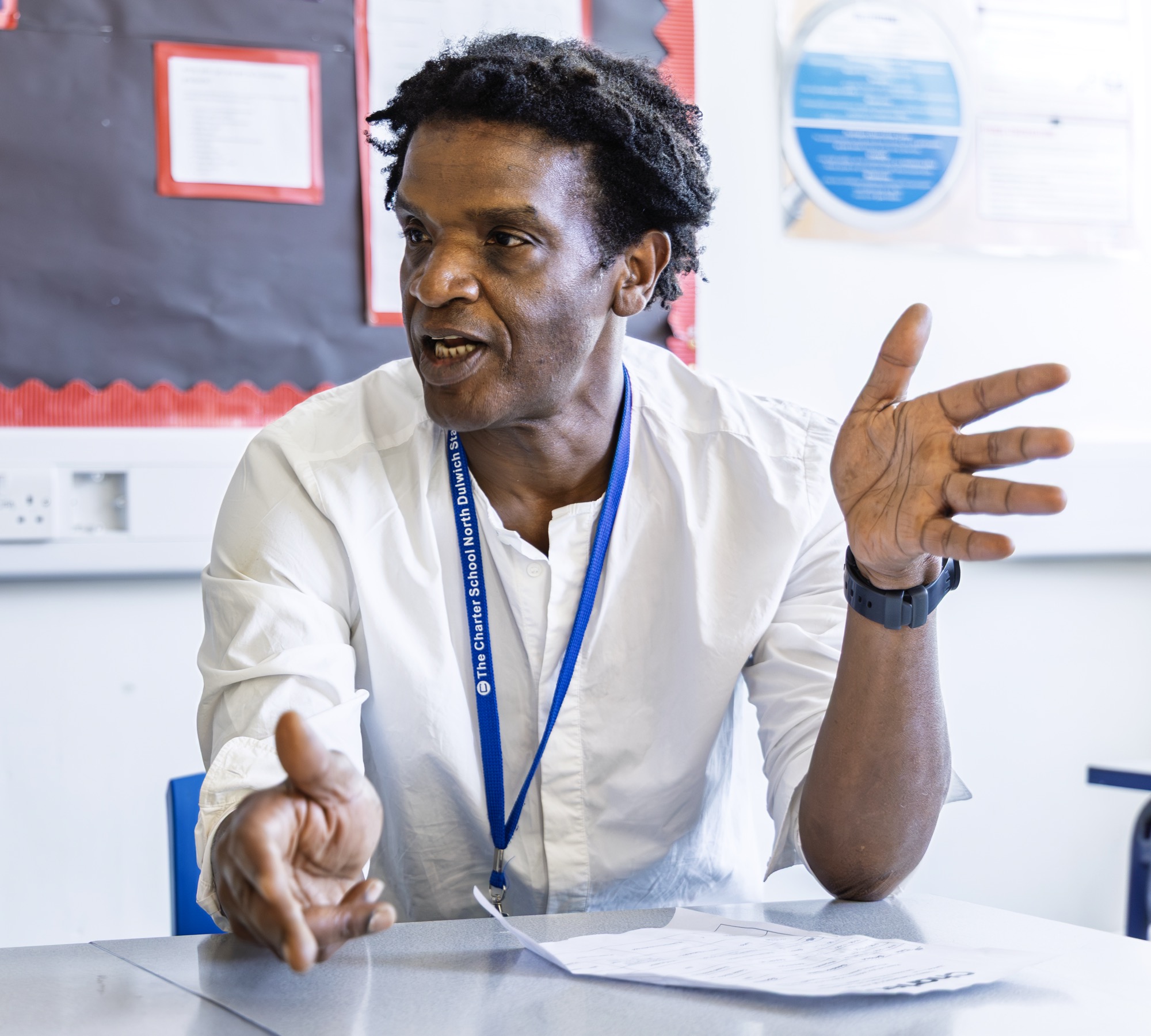 Man with dark curly hair wearing a white shirt and a blue lanyard, sitting at a table in a classroom, speaking and gesturing with both hands, with papers on the table and a bulletin board in the background.