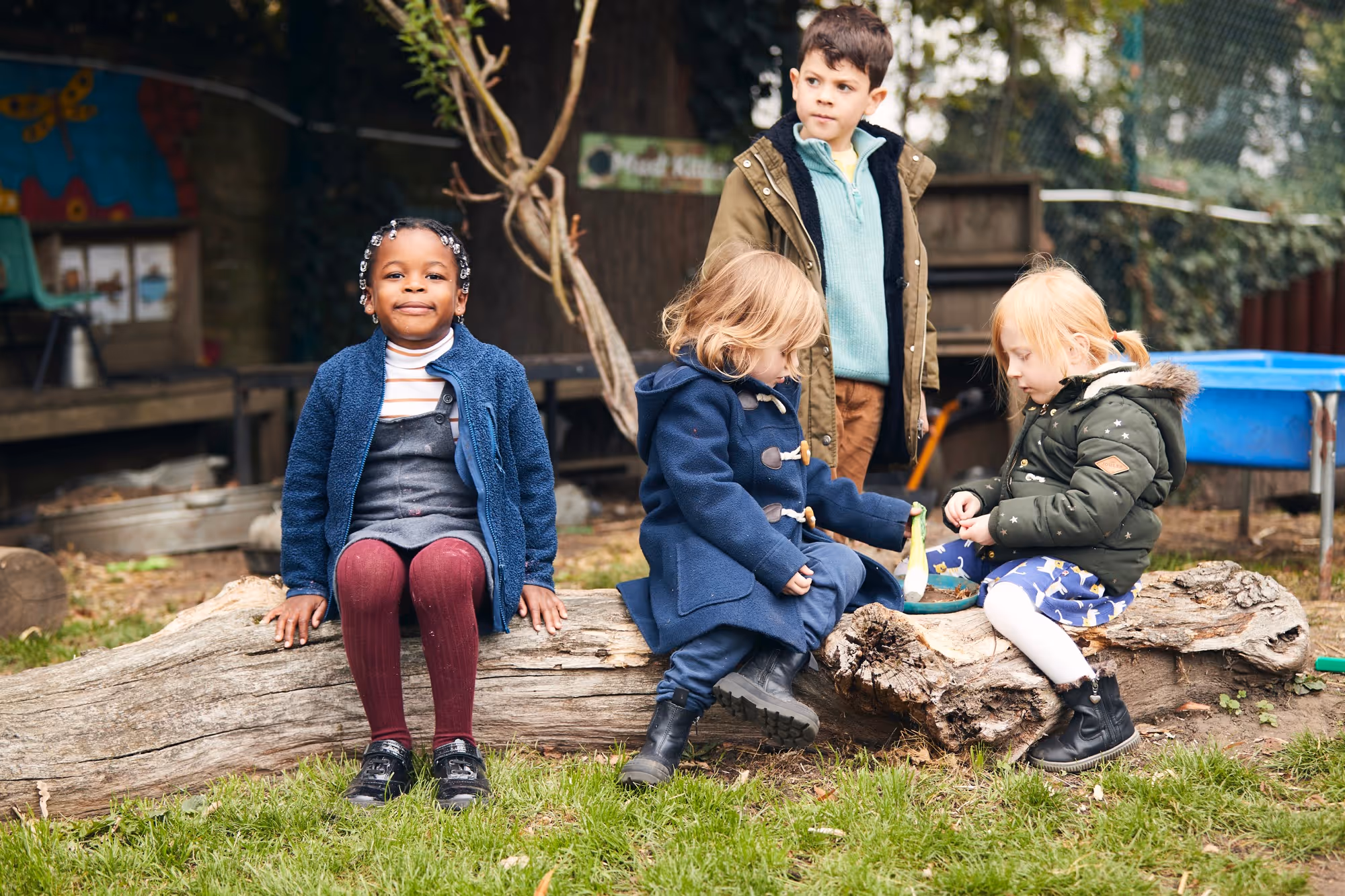 Four young children outdoors playing and sitting on large wooden logs in a grassy area, surrounded by trees and outdoor play equipment.