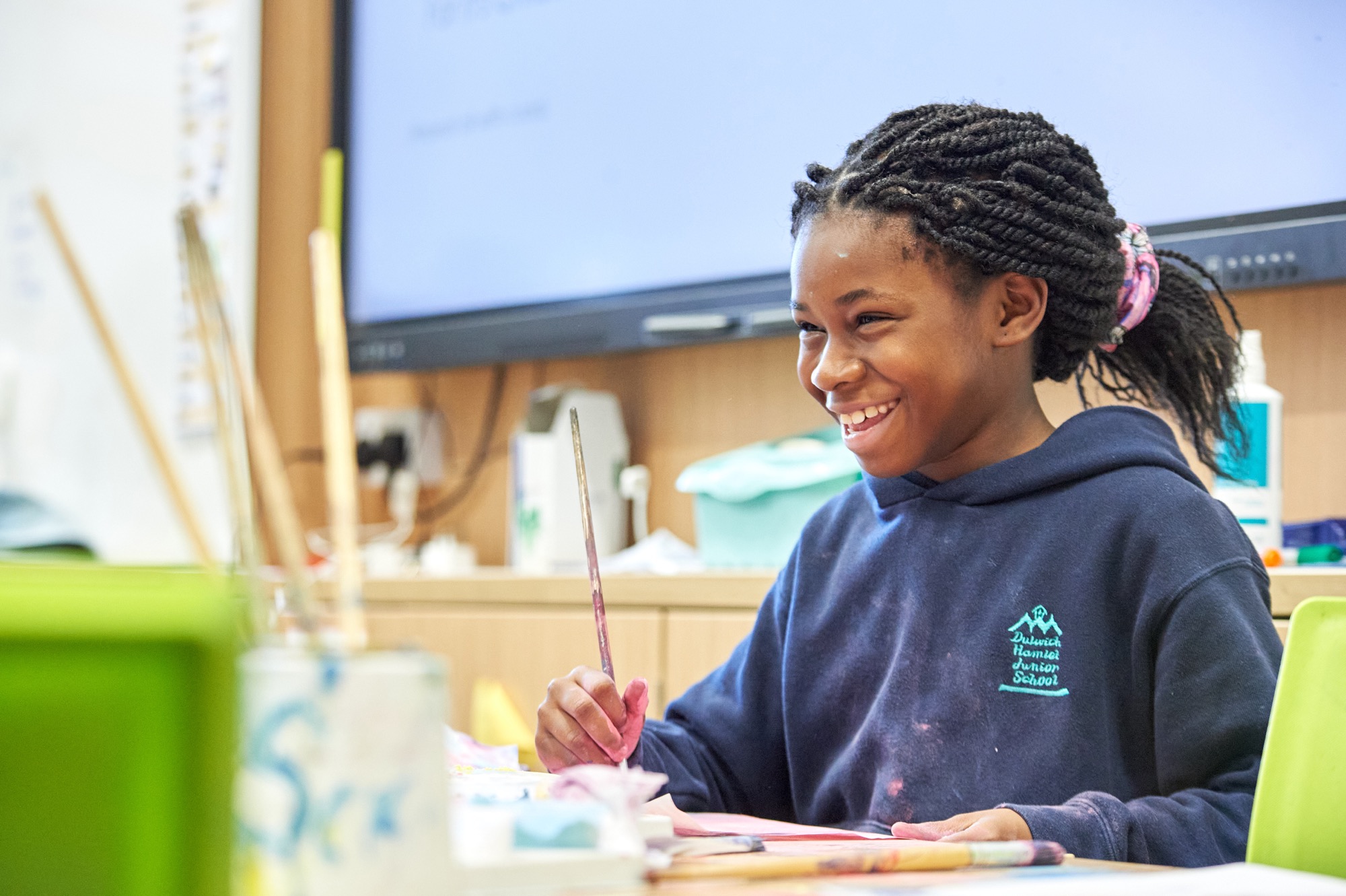 Smiling girl with braided hair wearing a navy blue Dulwich Hamlet Junior School hoodie, holding a paintbrush in a classroom art setting.