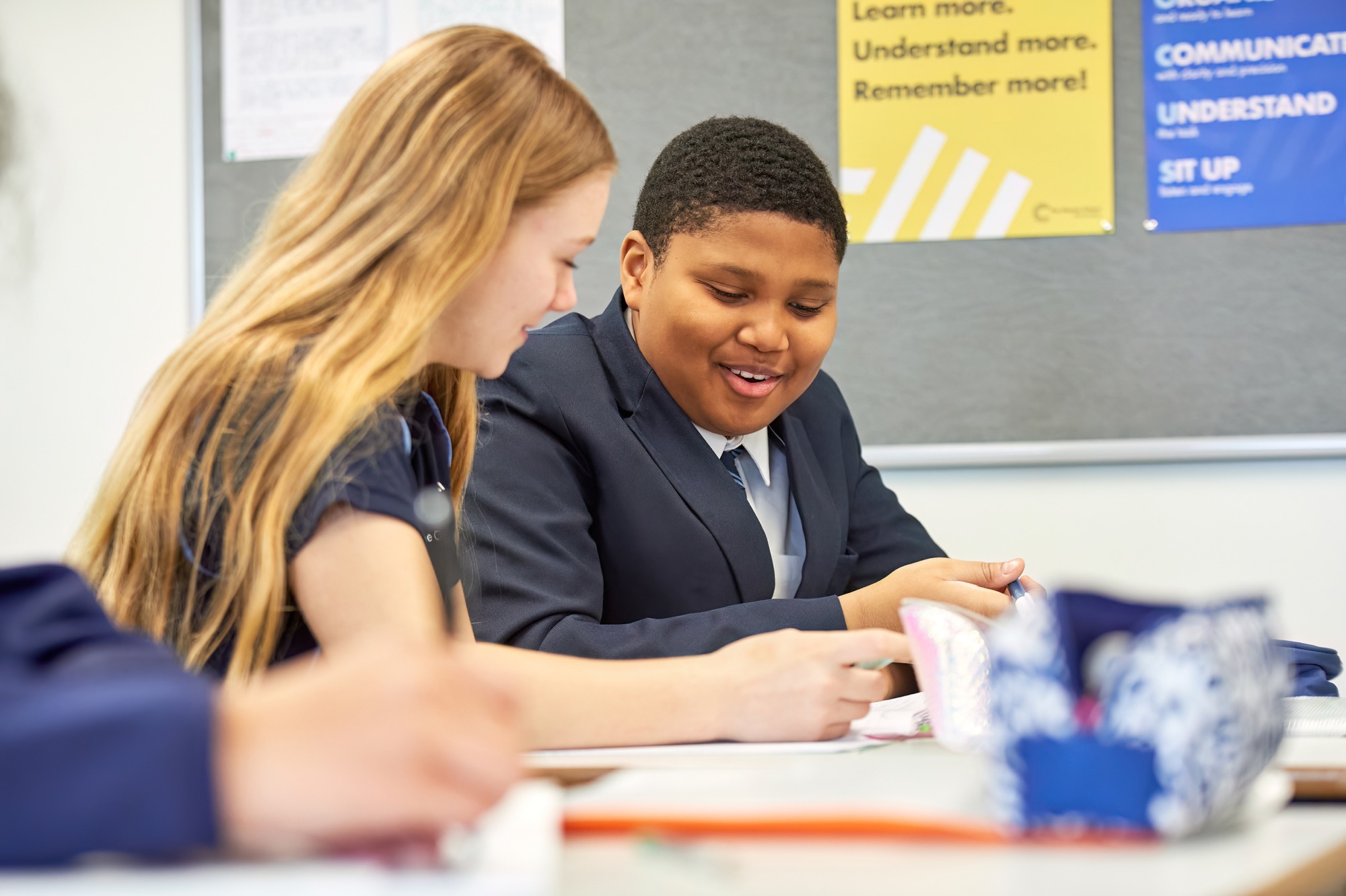 Two students in school uniforms sit at a desk in a classroom working together with focus, smiling and looking at materials on the table.
