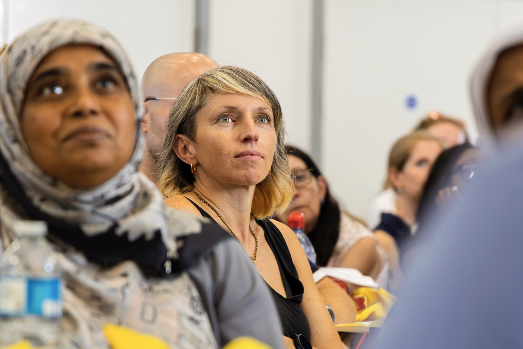 A diverse group of people attentively listening at a conference or seminar, with a focus on a woman with short blonde hair wearing a black top.