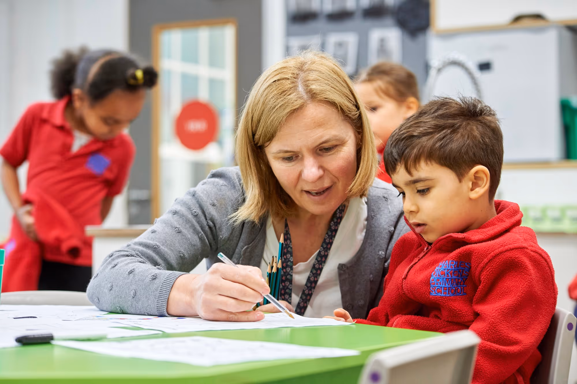 Teacher helping a young boy with schoolwork at a green table in a classroom, while other children are in the background.