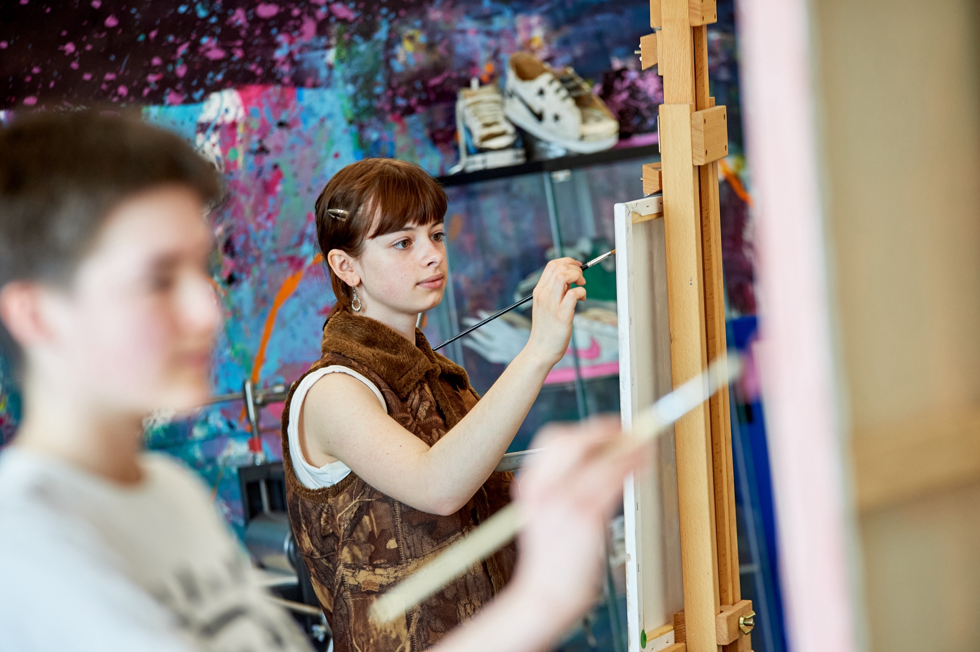 Young woman with short brown hair painting on a canvas set on an easel in a colorful art studio, with a blurred person painting in the foreground.