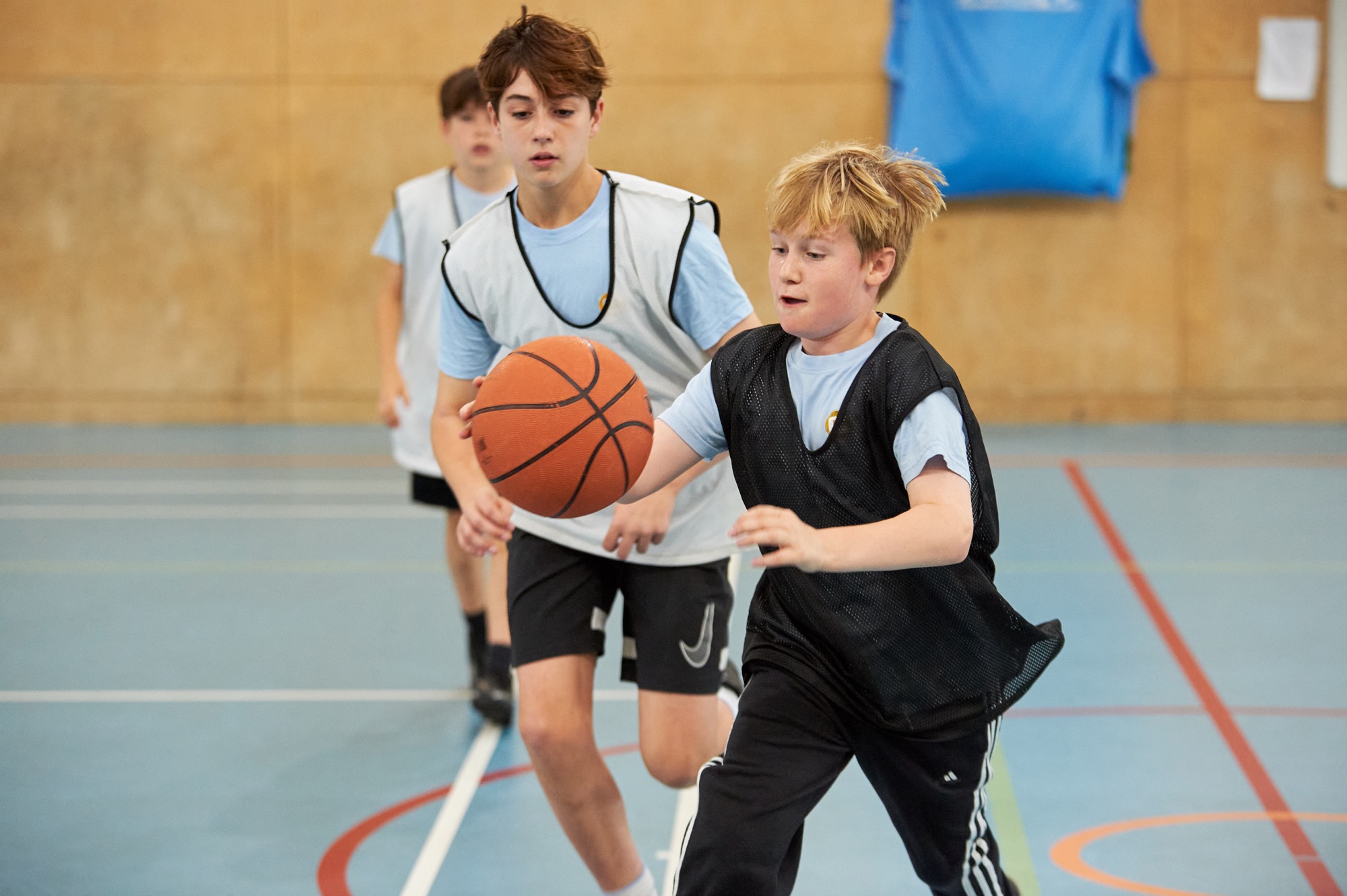 Three boys playing basketball indoors, with one boy in a black vest dribbling the ball while a boy in a white vest closely follows him.
