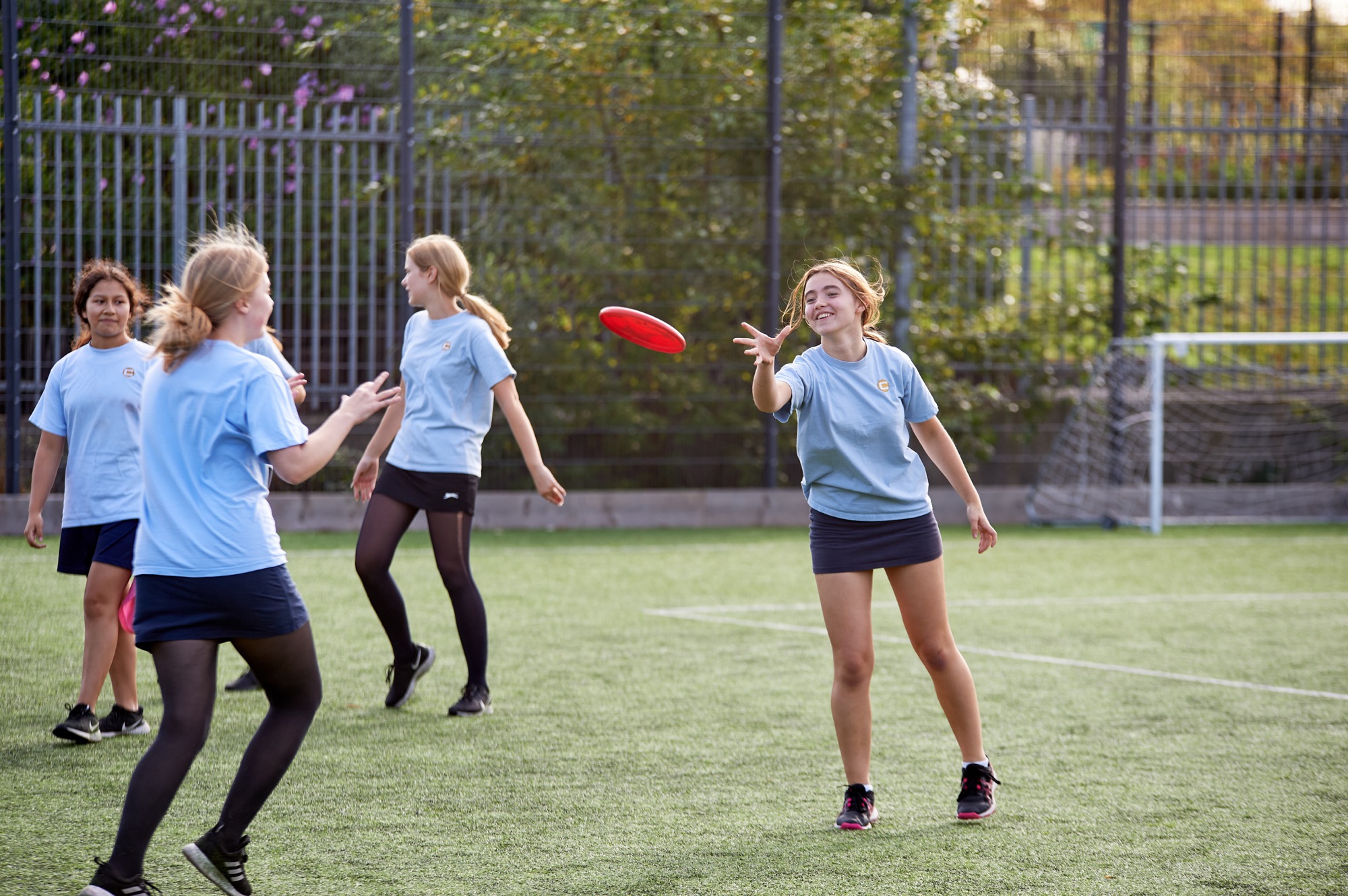 Four girls wearing light blue shirts and dark skirts or shorts playing frisbee on a green soccer field with a goal and fence in the background.