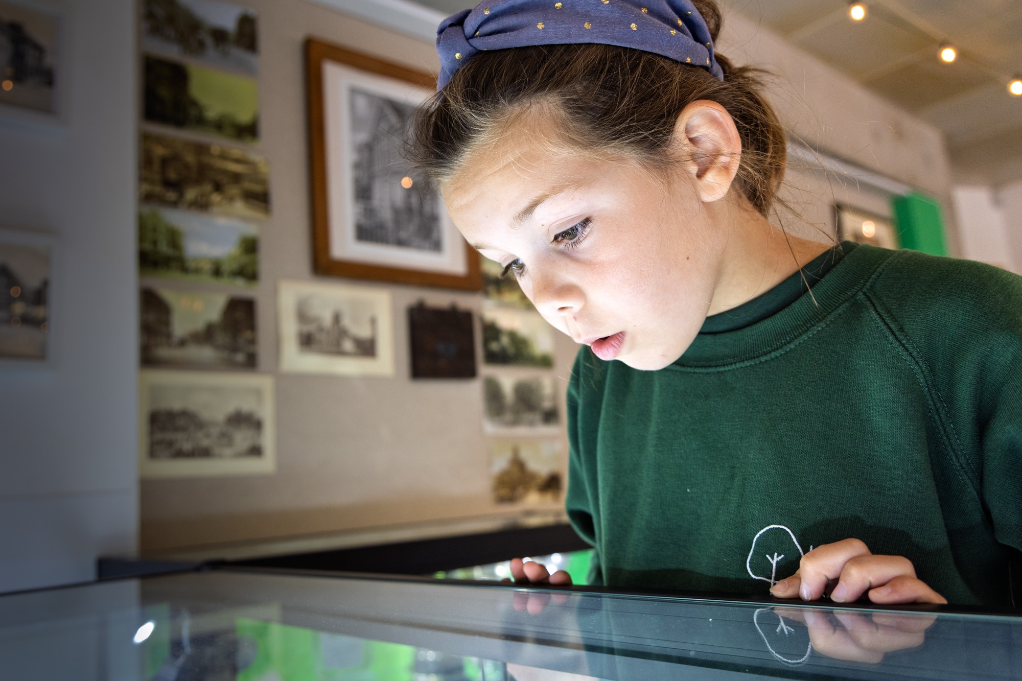 A young girl with a blue headband and green shirt intently looks at an illuminated display table in a museum or gallery setting with framed pictures on the wall behind her.