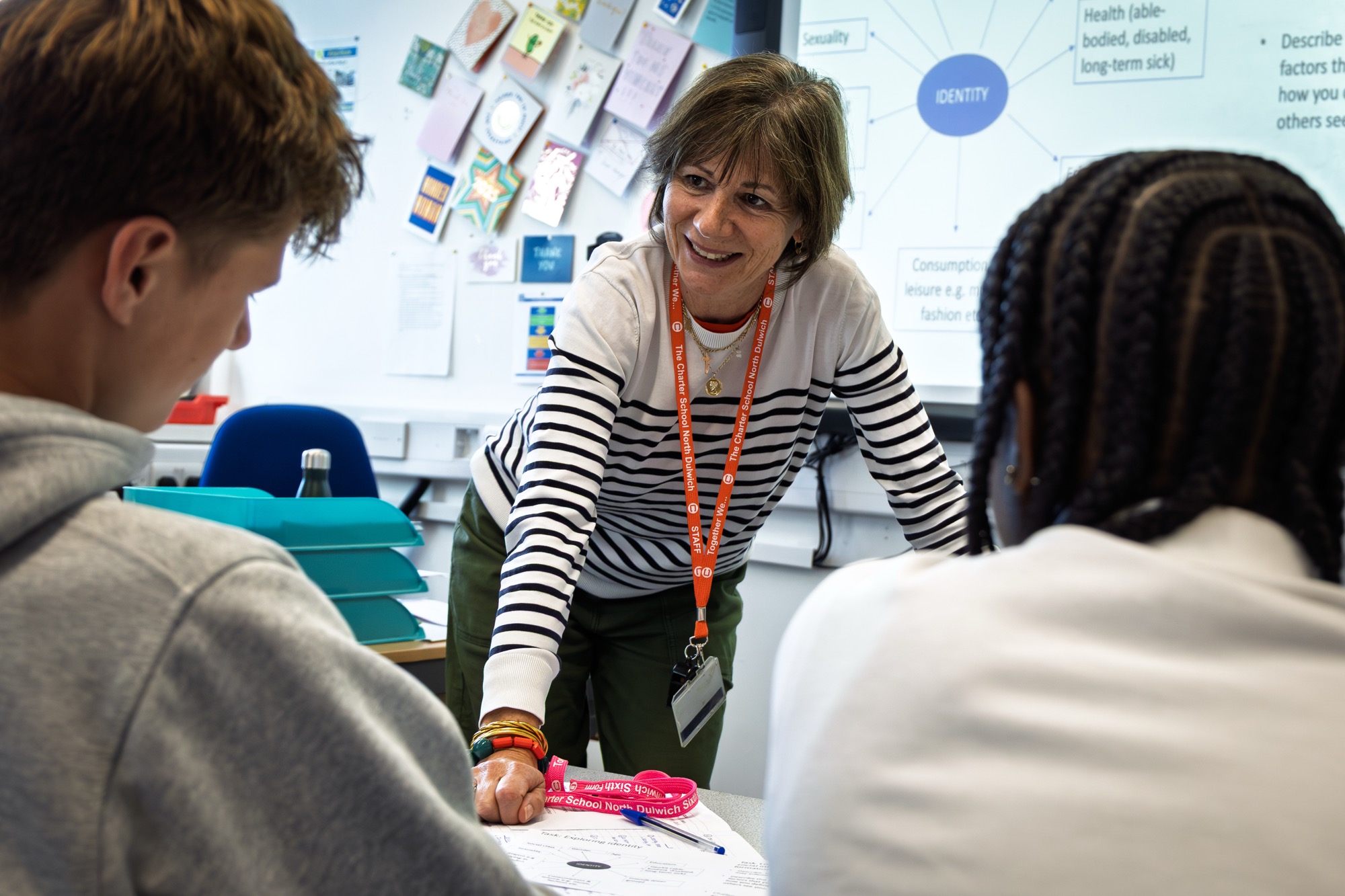 Teacher leaning on desk engaging with two students in a classroom with educational posters on wall behind.