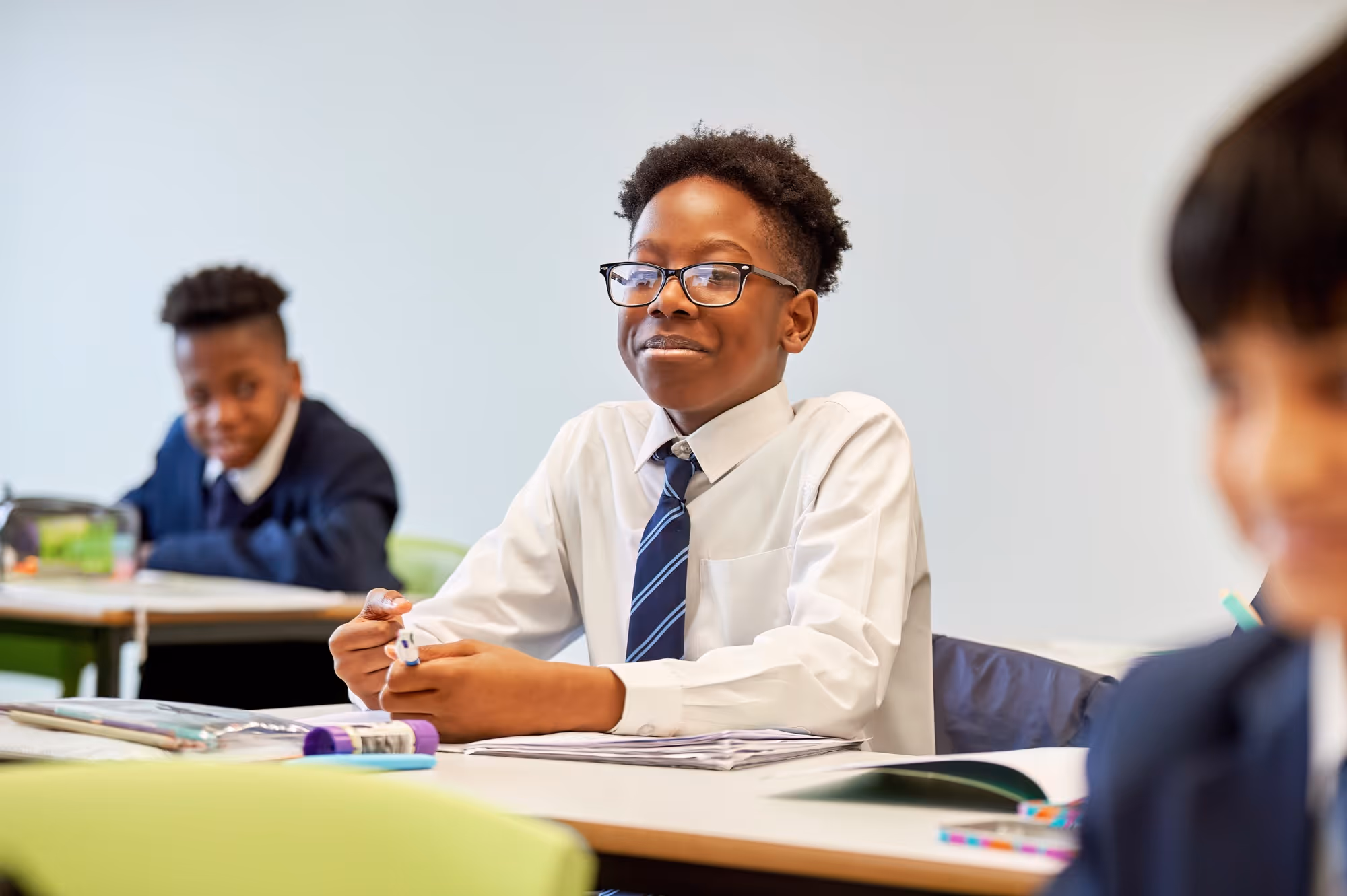Young student in a white shirt and tie sitting at a desk in a classroom with other students.