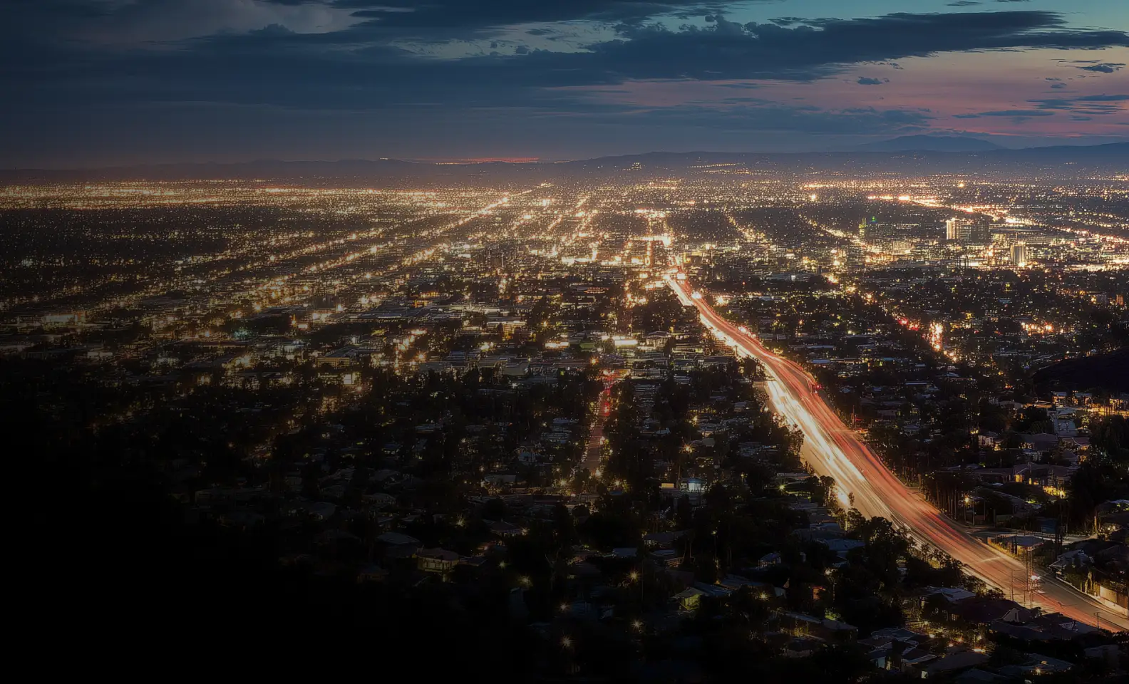 Cityscape at dusk with illuminated streets and a busy highway extending into the distance.
