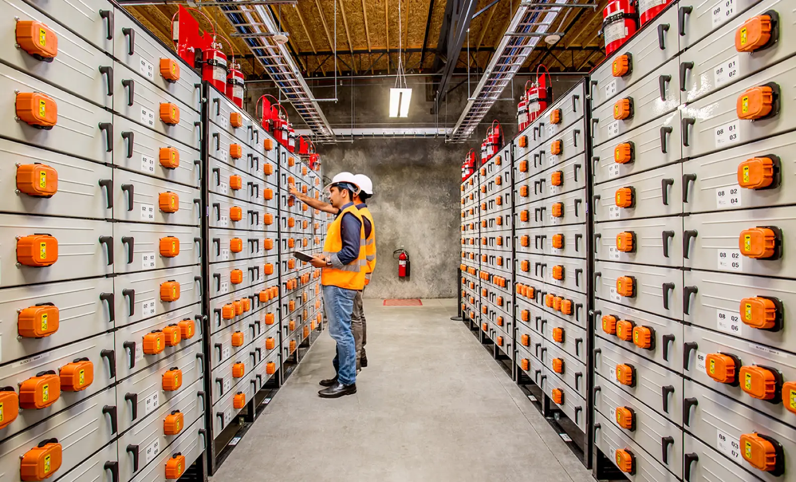Two workers in safety vests and helmets inspecting industrial electrical panels with orange switches in a utility room.