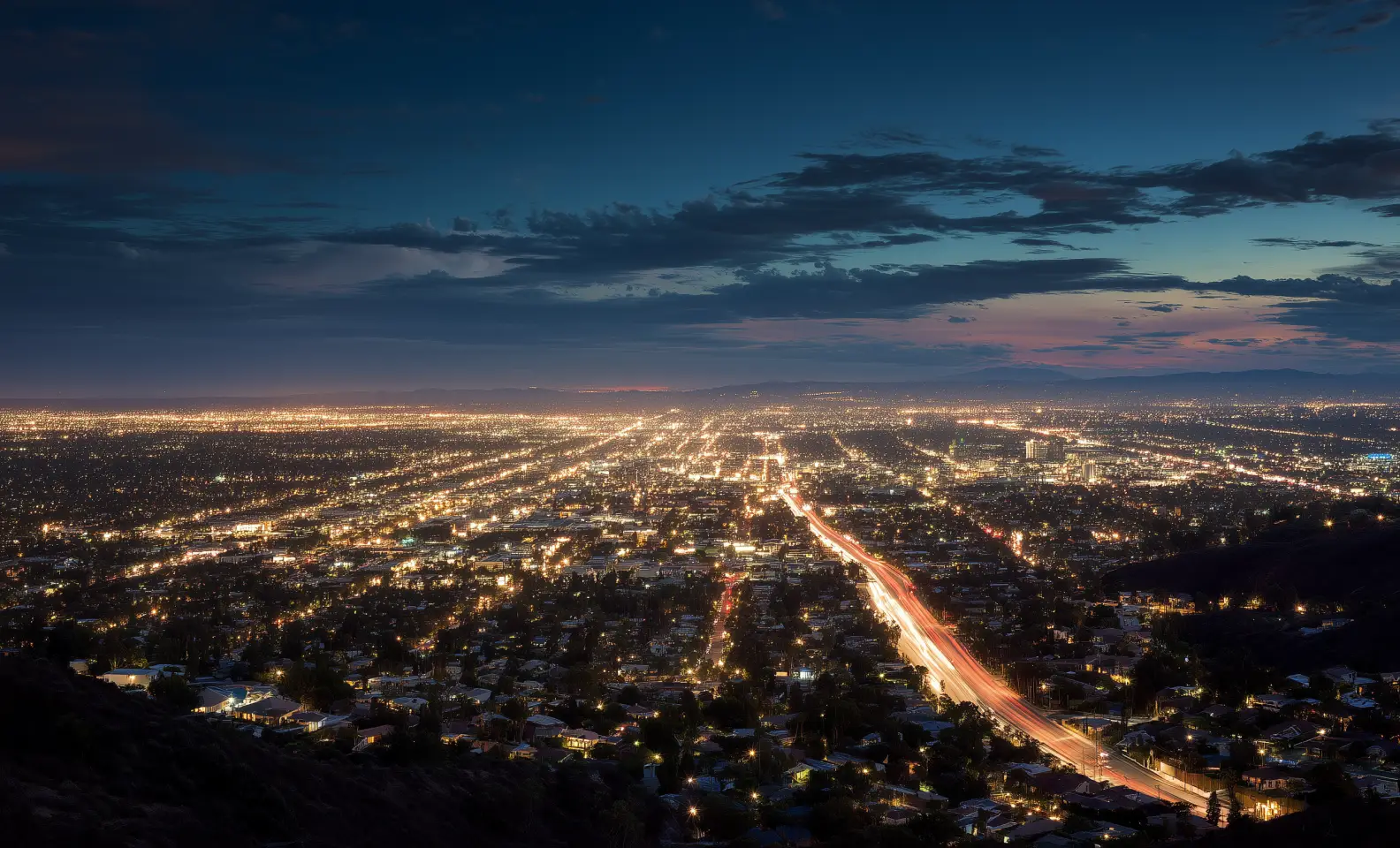 Nighttime cityscape with illuminated streets and buildings stretching into the distance under a partly cloudy sky.