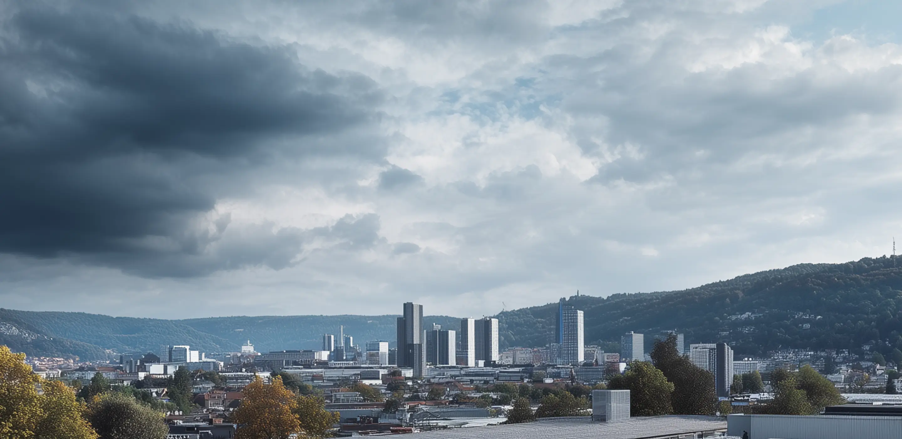 City skyline with tall buildings under a cloudy sky with dark clouds on the left and hills in the background.