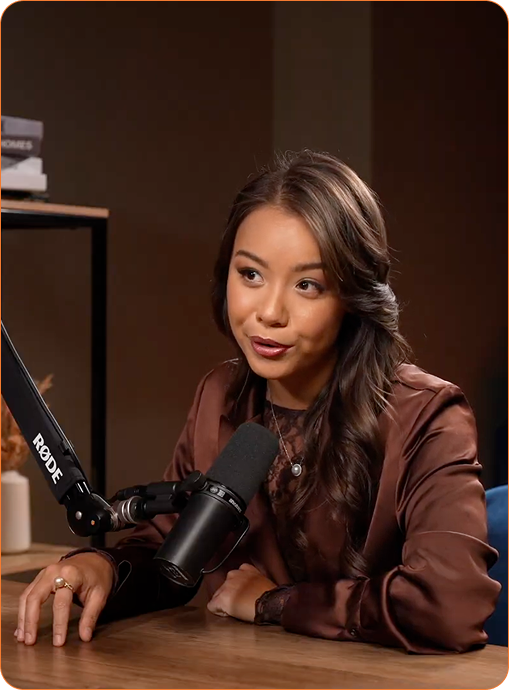 Woman with long dark hair speaking into a Rode microphone at a table in a recording studio.