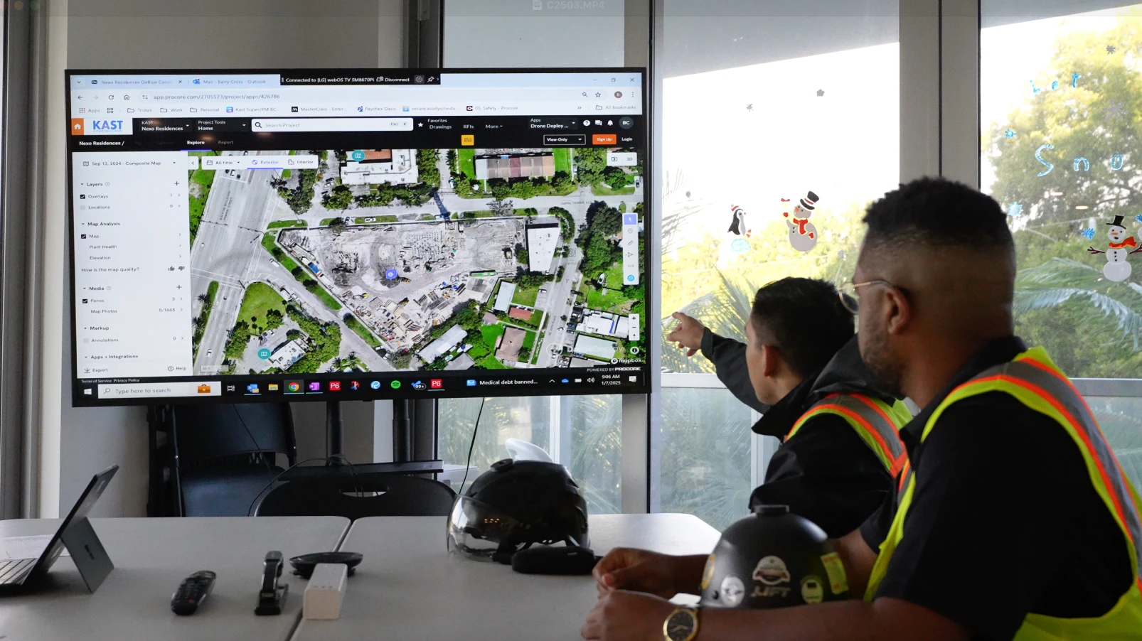 Two construction professionals in safety vests reviewing aerial site imagery on a large screen using DroneDeploy software during a project meeting in a conference room.