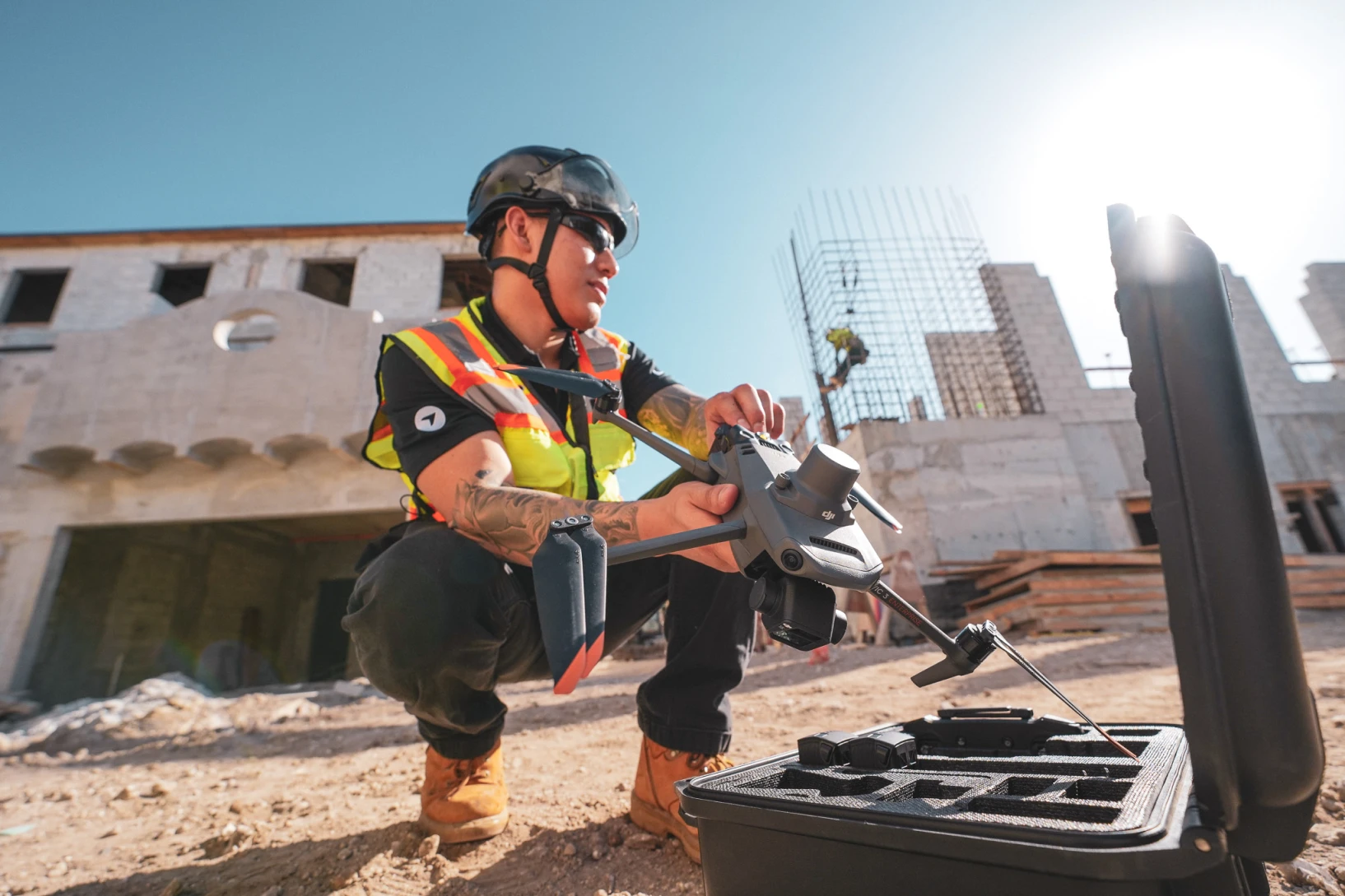 Construction worker in safety gear preparing a drone for flight on an active jobsite, with rebar columns and partially built concrete structures in the background.