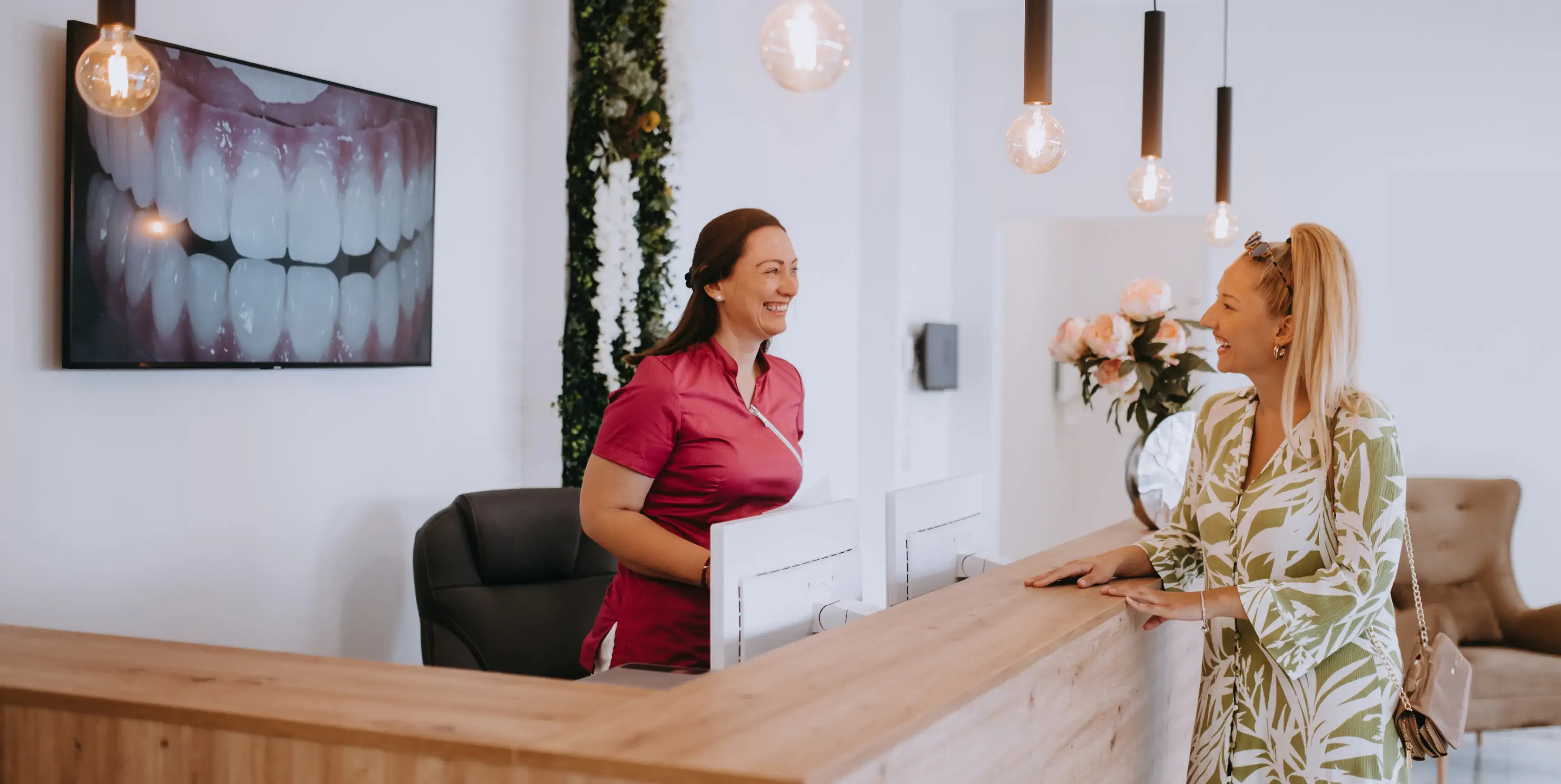 Receptionist in red uniform smiling and talking with a female patient at a dental clinic reception desk.