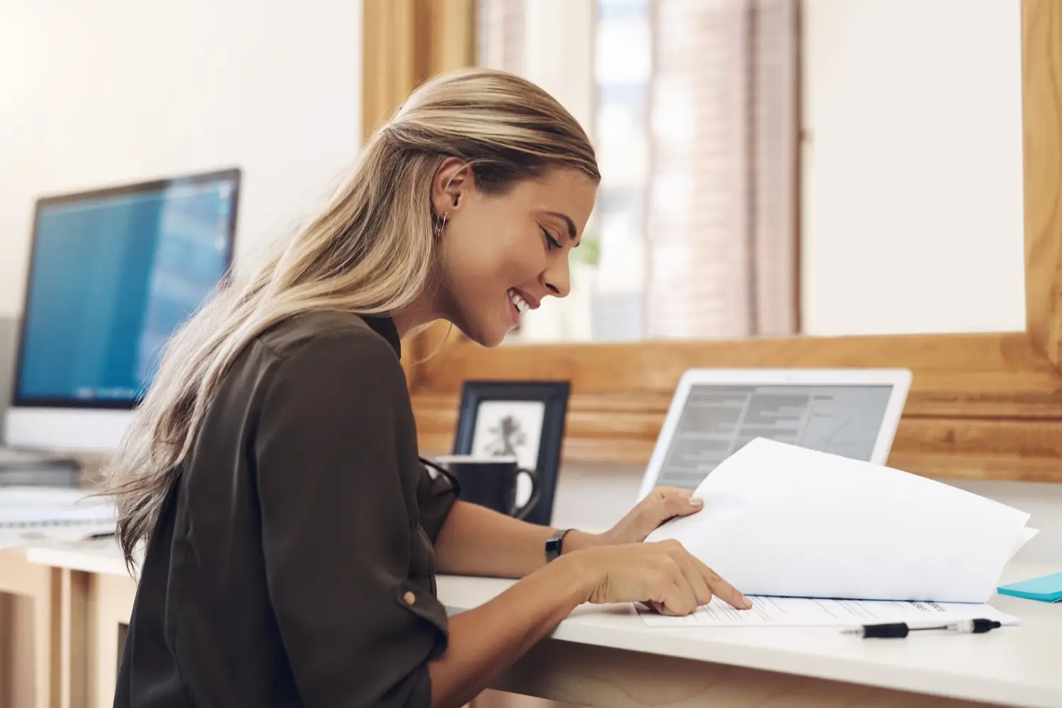 Smiling woman reading documents at a desk with a laptop and pen nearby.