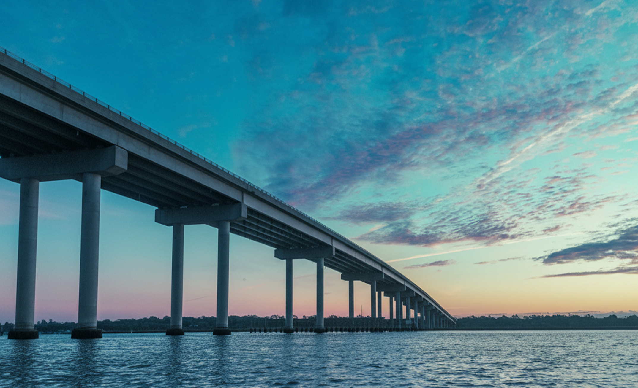 Long concrete bridge spanning over water under a blue and purple cloudy sky at sunset.