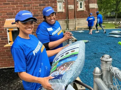 Two smiling volunteers in blue Progressive shirts holding a large bag of blue rubber mulch.