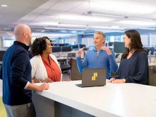 Four coworkers chatting and smiling around a white table with a laptop in an open office.