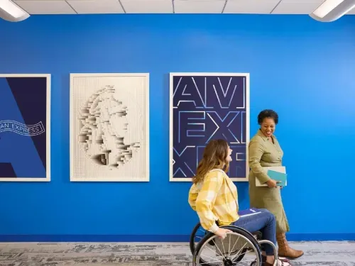 Two women smiling and walking in an office, one using a wheelchair, with blue walls and framed art.