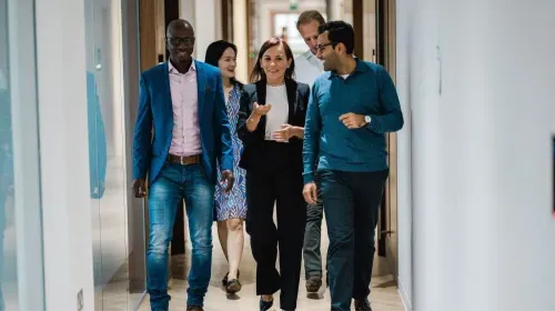 Five professionals walking and talking together in a bright office hallway.