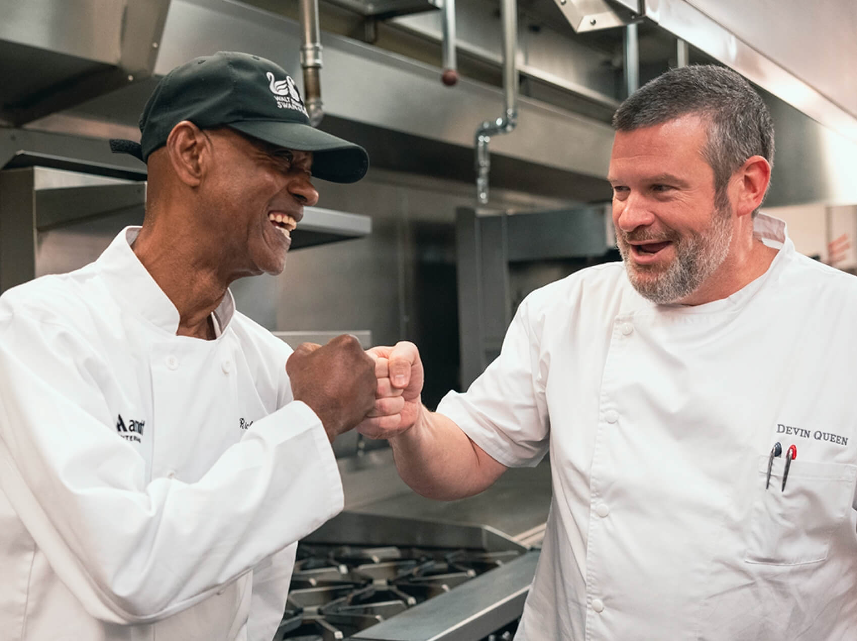 Two Marriott chefs in a commercial kitchen smile and exchange a fist bump while standing beside industrial cooking equipment, highlighting teamwork, collaboration, and a positive workplace culture.