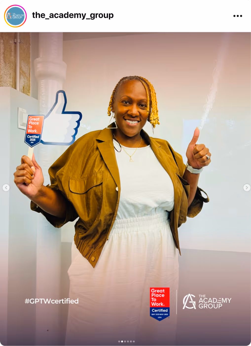 Person holding a thumbs‑up sign beside a Great Place To Work Certification badge in an indoor setting.