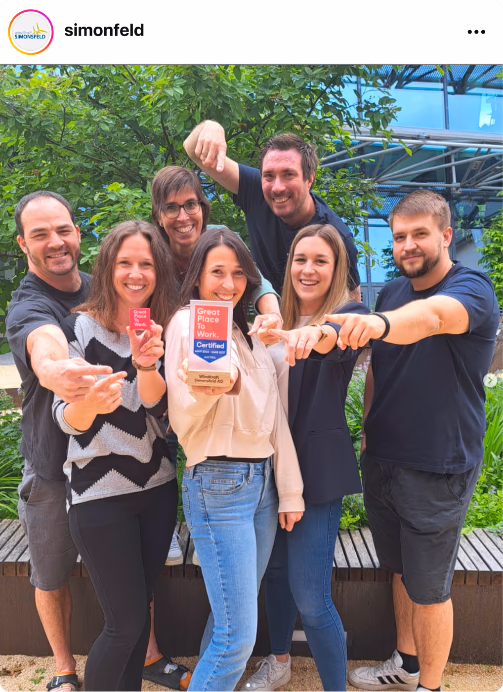Group of colleagues pointing toward the camera while holding a Great Place To Work Certification badge outdoors.