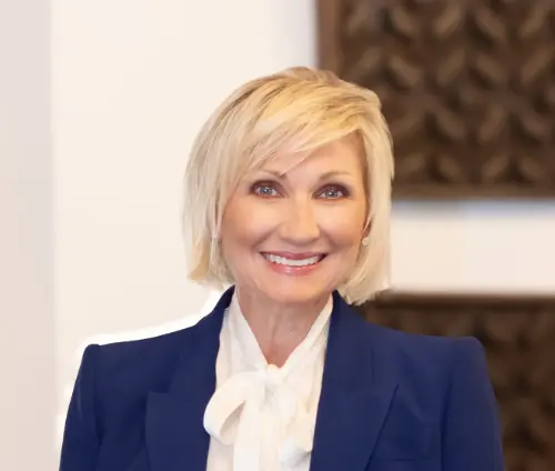 Smiling woman with short blonde hair wearing a blue blazer and white blouse bow tie.
