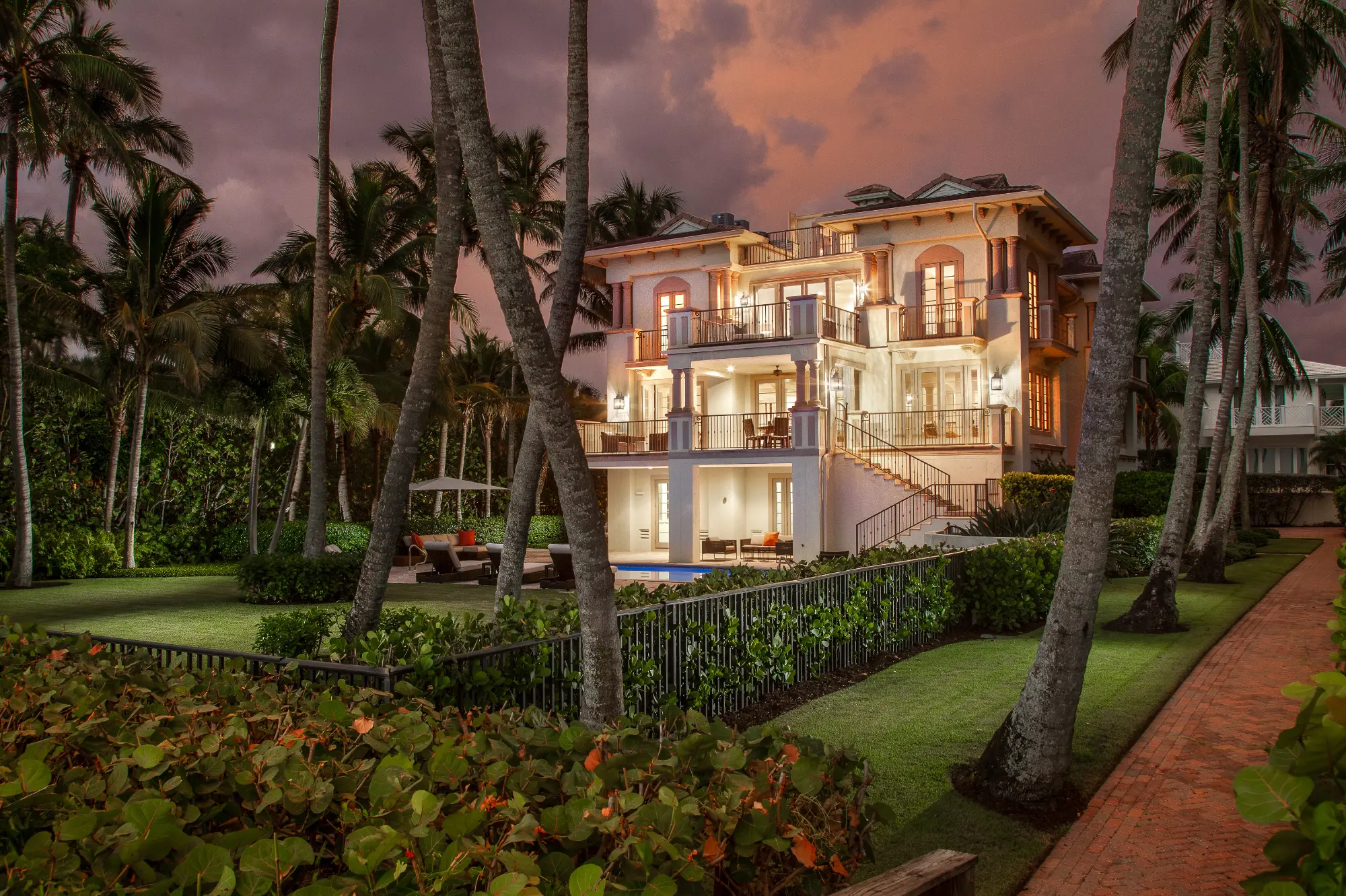 Large illuminated mansion with balconies, surrounded by palm trees and lush gardens at dusk.