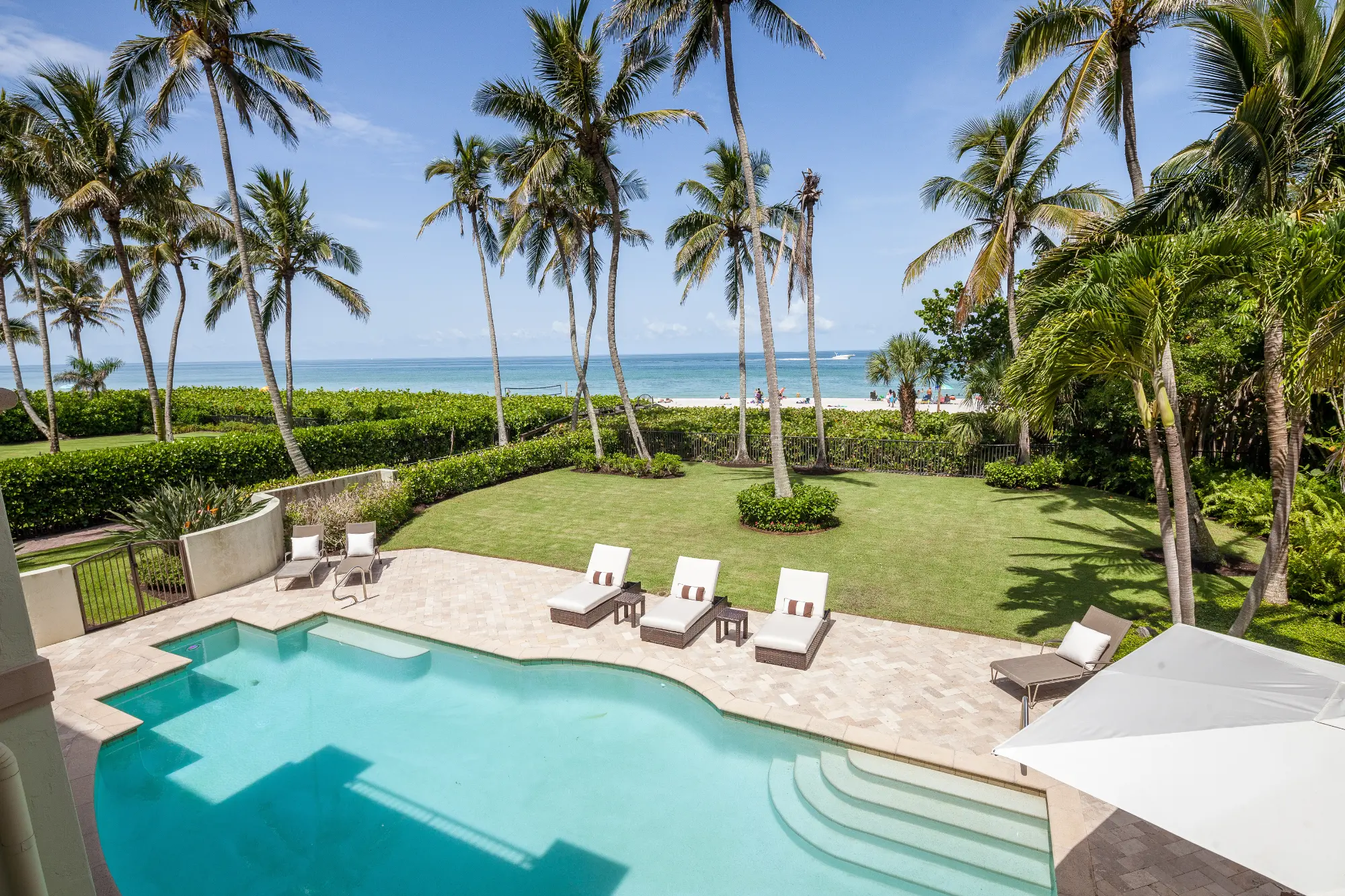 Naples, Florida oceanfront backyard with pool, lounge chairs, palm trees, and green lawn under a clear sky.