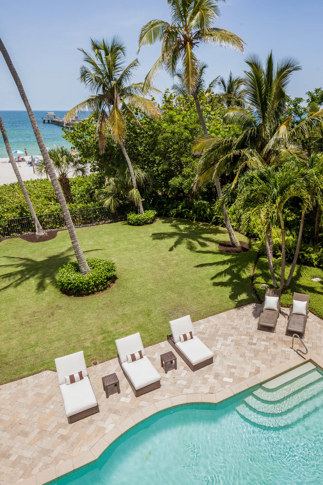 Outdoor pool with lounge chairs, palm trees, green lawn, and a beach with ocean and pier in background.