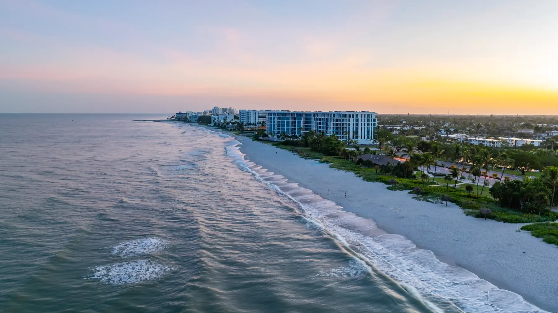 Coastal beachfront with gentle waves, white sand, palm trees, buildings, and a colorful sunset sky.