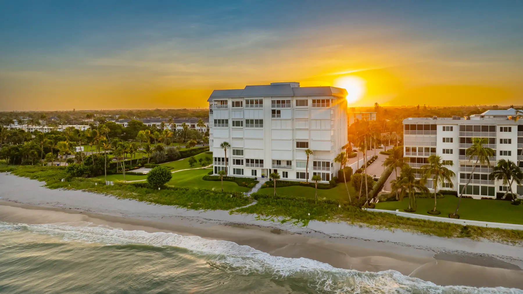 Beachfront white building at sunset with waves, sandy shore, palm trees, and lush greenery.