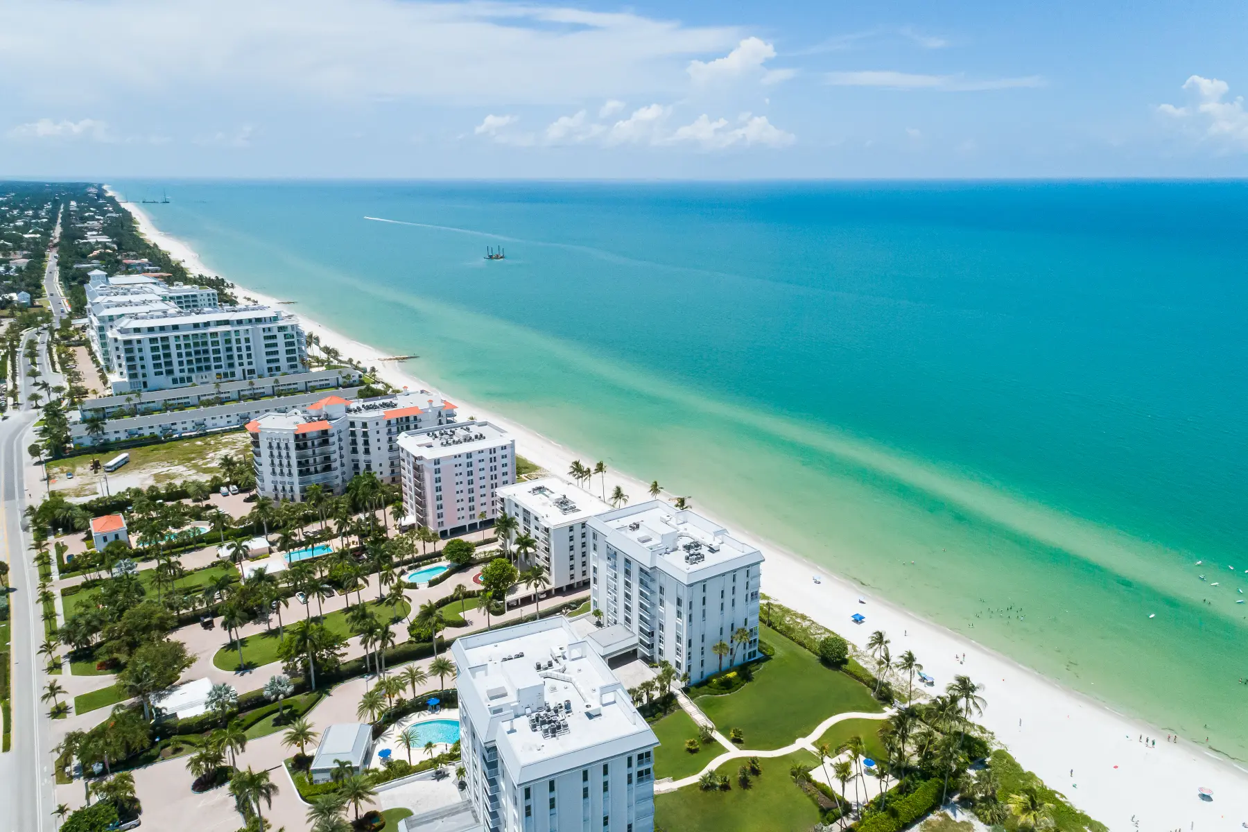 Aerial view of beachfront buildings with white sand beach, turquoise ocean, and a partly cloudy sky.